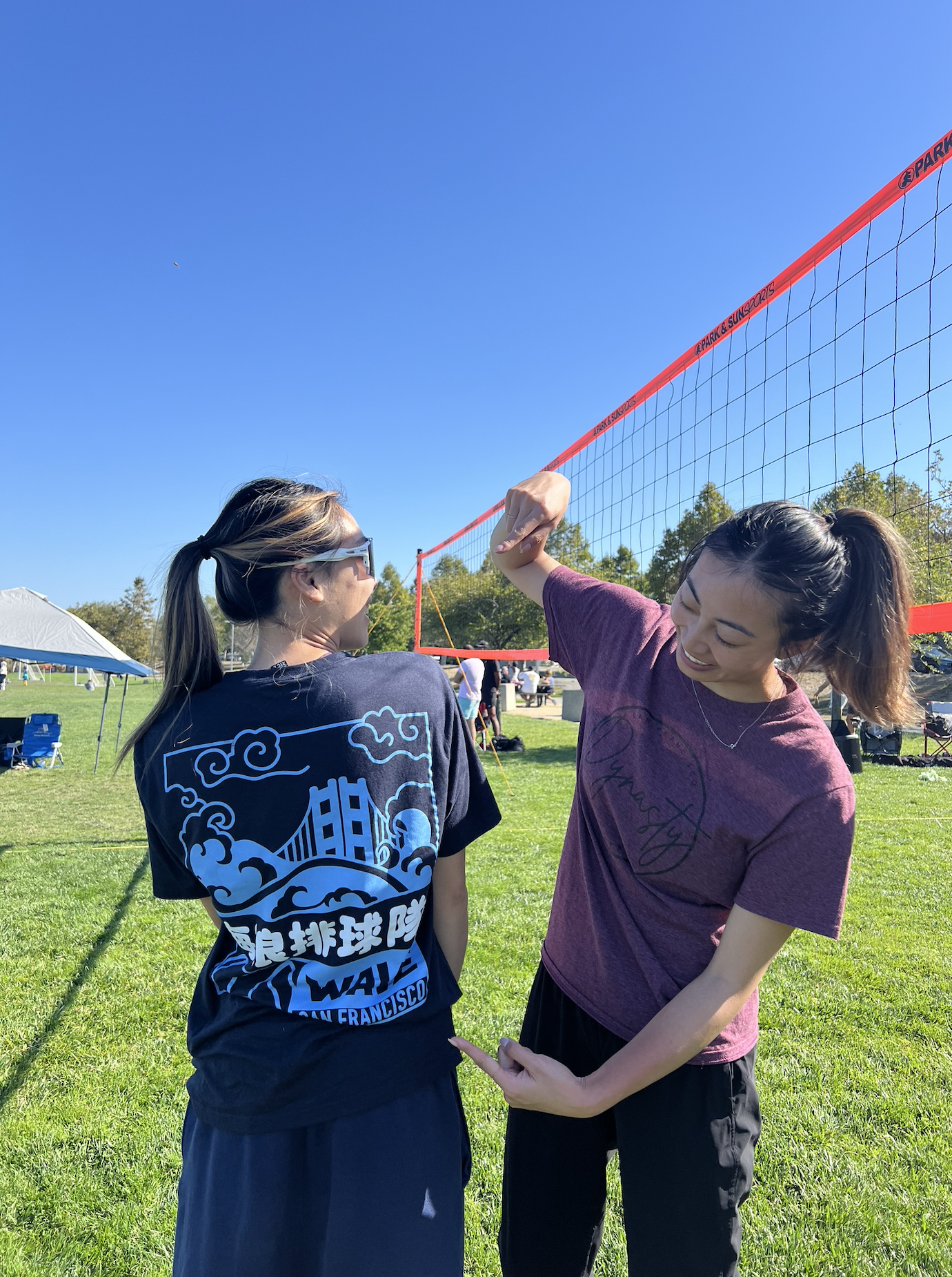 Two women are smiling and talking near a volleyball net on a grassy field with clear blue skies, trees, and tents in the background.