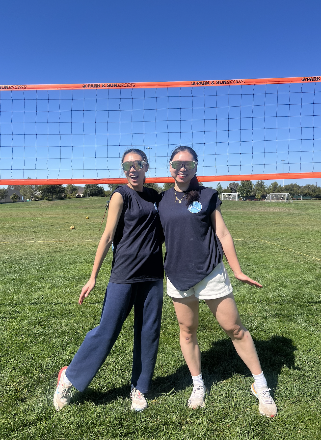 Two women standing on a grassy field in front of a volleyball net, smiling and posing. Both are wearing sunglasses and black shirts, with a blue sport logo, and athletic pants or shorts. The sky is clear and blue, and there are soccer nets visible in