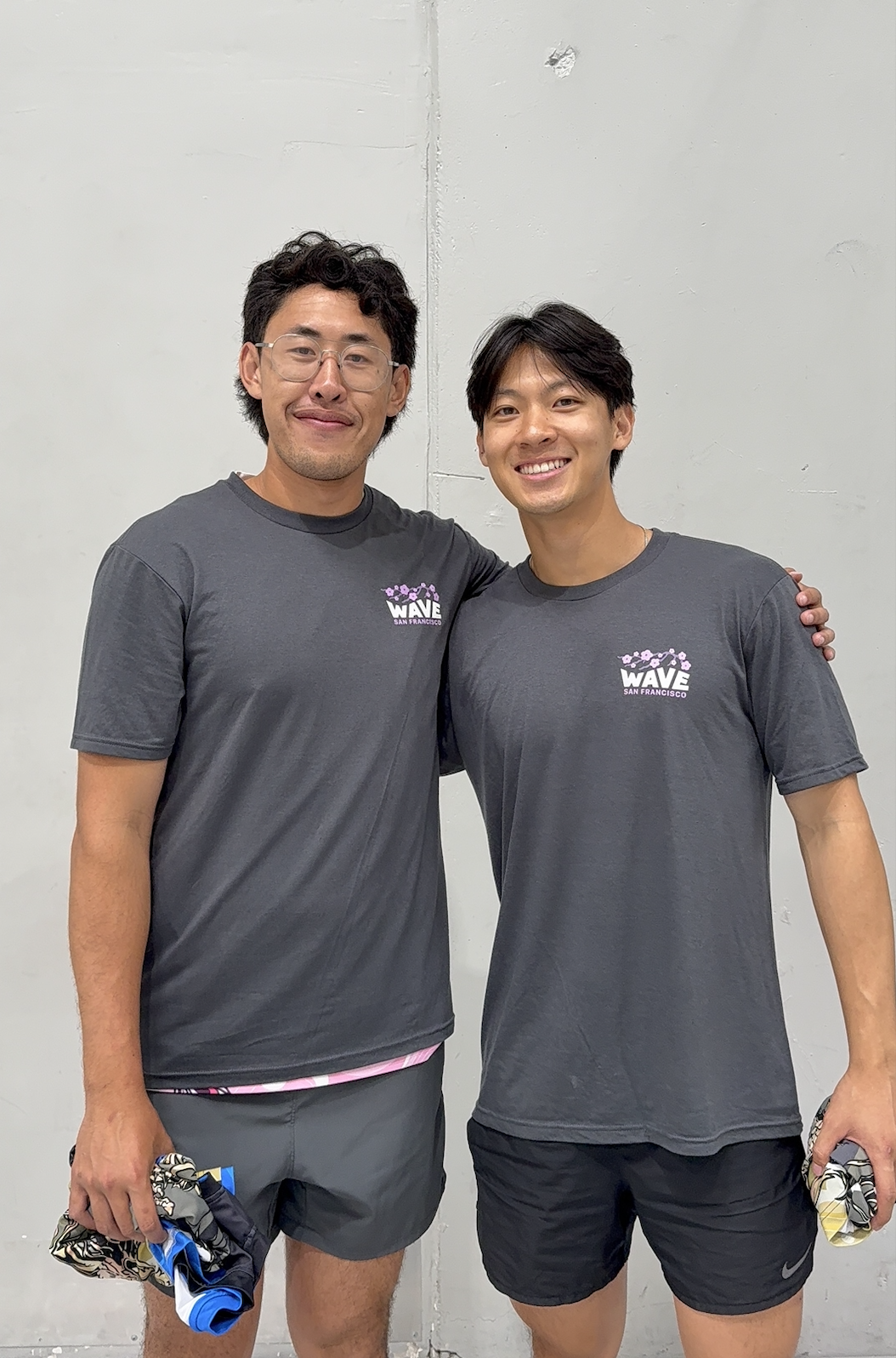 Two young men in dark gray t-shirts with 'Wave San Francisco' logo, smiling, standing close against a plain wall.