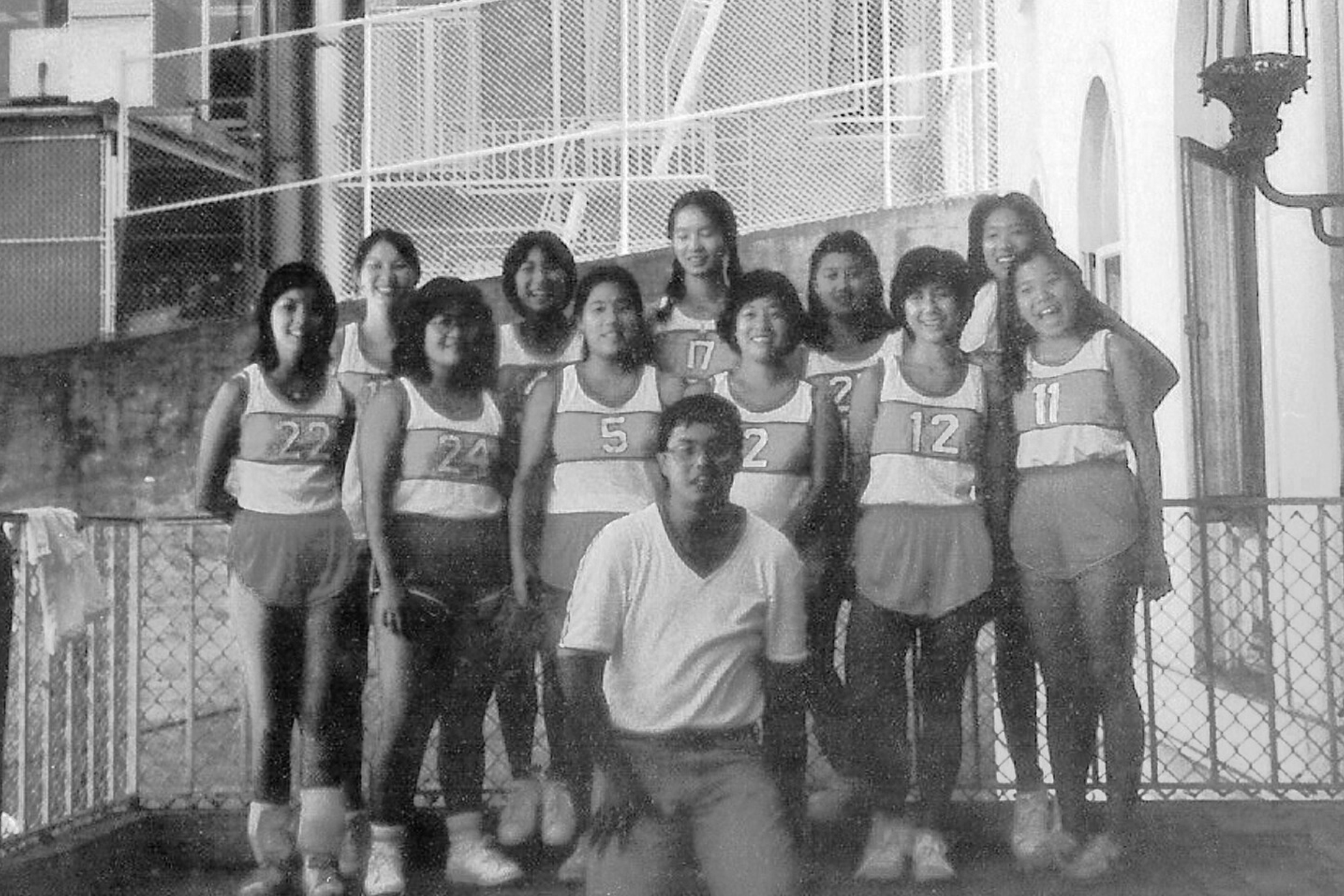 Group of young women and one young man posing for a photo outside a sports facility with a chain-link fence, all wearing athletic uniforms with numbers.