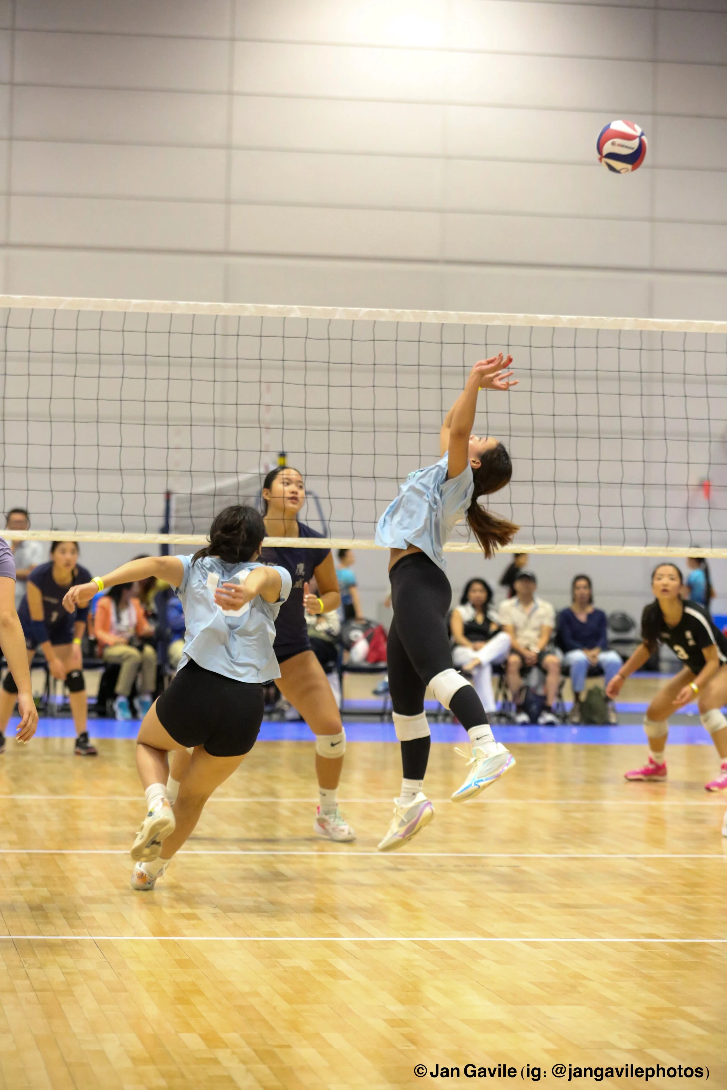 Women volleyball players jumping to hit ball over net during indoor game with spectators in background.