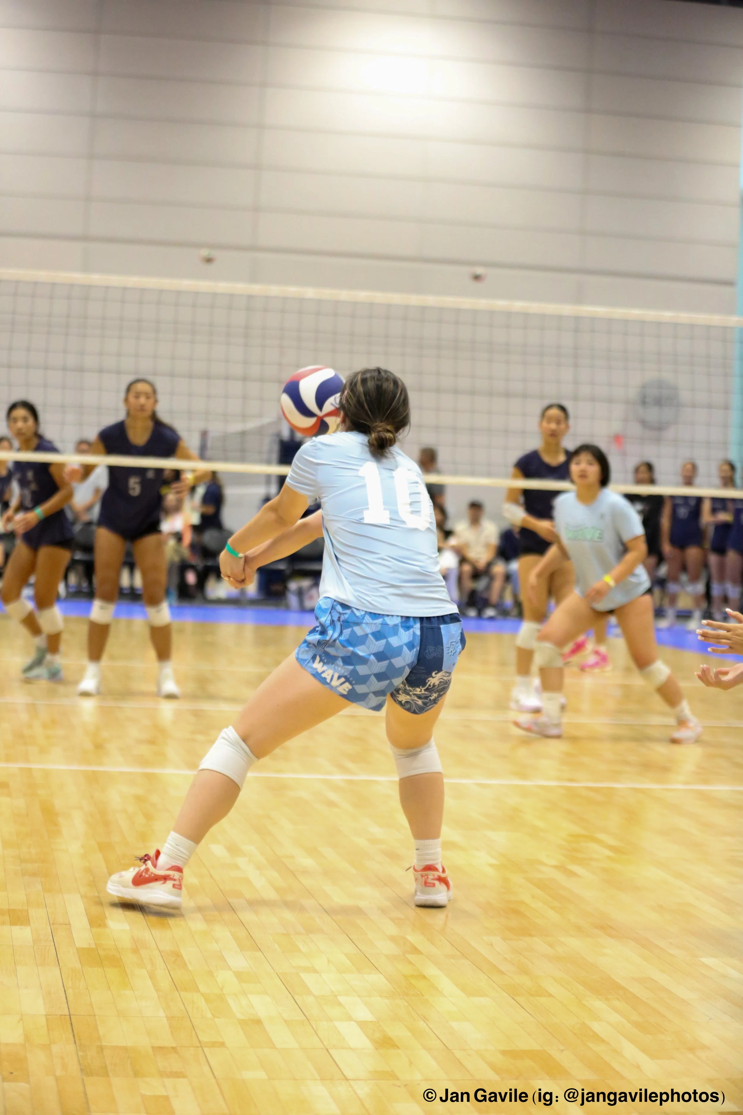 A volleyball game in an indoor gym with female players. The focus is on a player in a light blue uniform with the number 10, preparing to hit the volleyball, while opponents in dark blue uniforms are on the other side of the net.