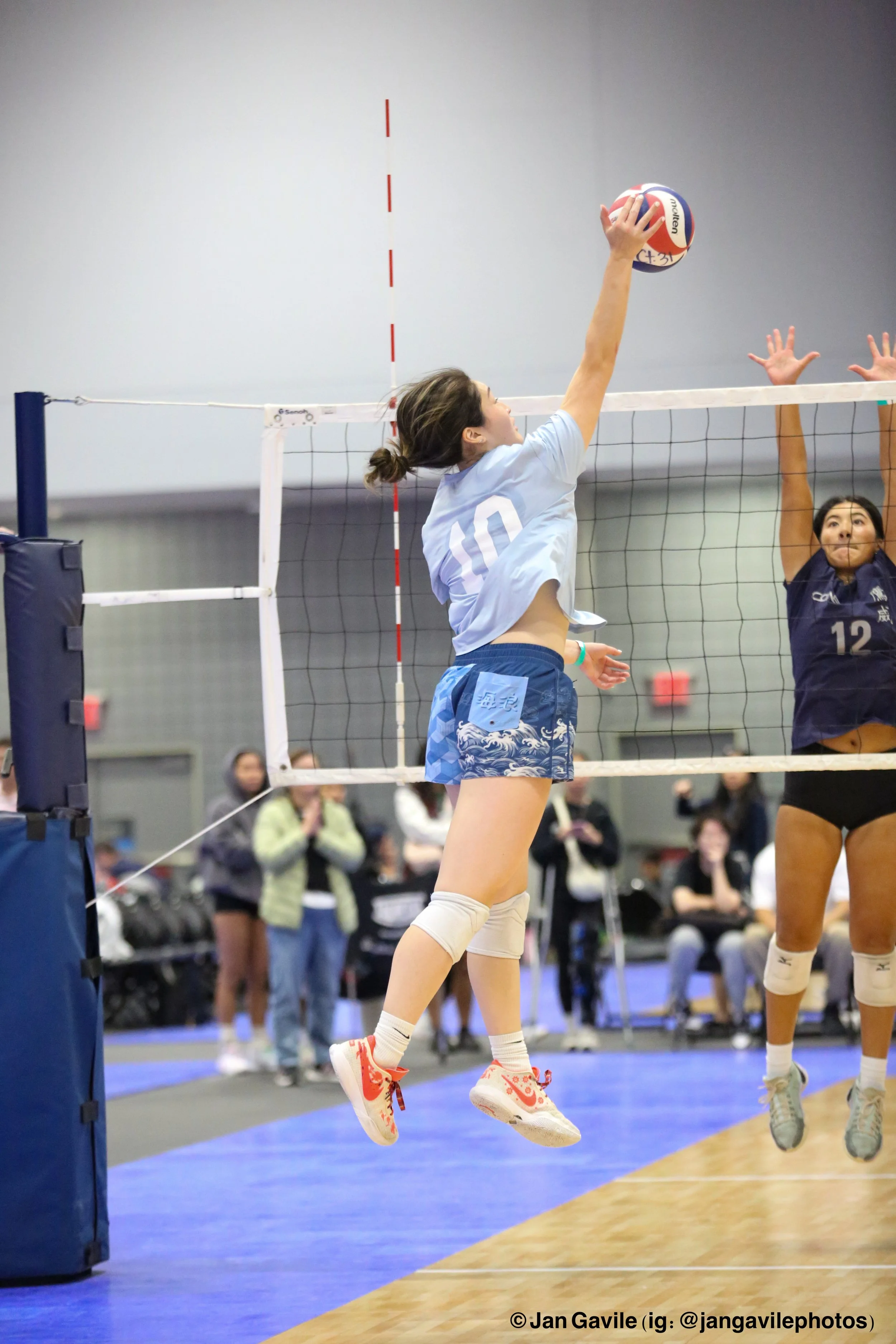 A female volleyball player with dark hair tied back, wearing a light blue jersey with the number 10, and patterned blue shorts, jumps to hit the ball during a game. She is on a volleyball court with a net in front of her, and there are spectators wat