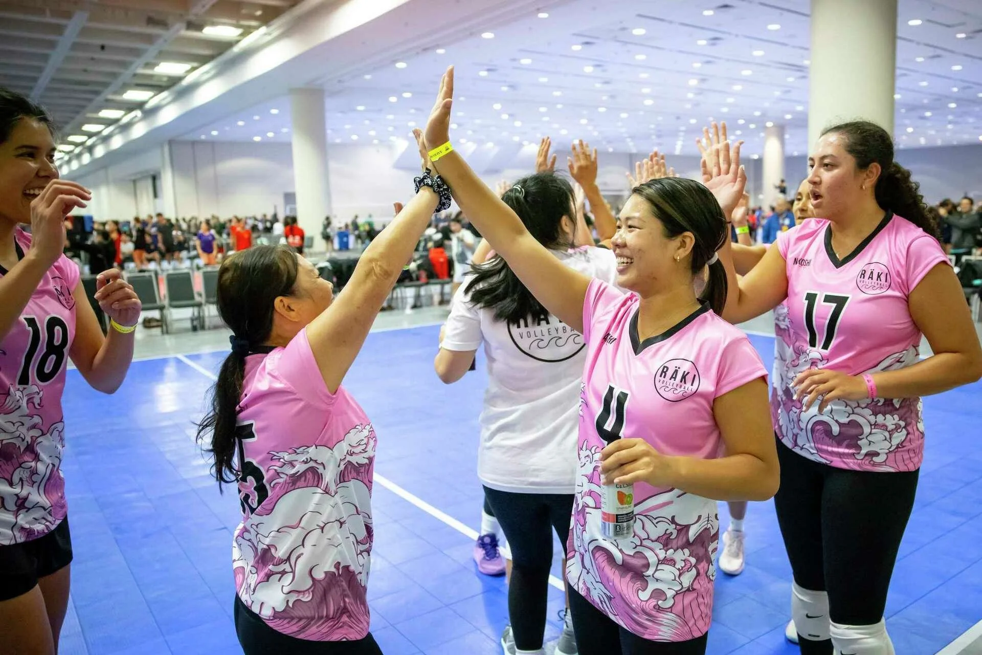 Group of female volleyball players wearing pink uniforms celebrating in an indoor sports arena.