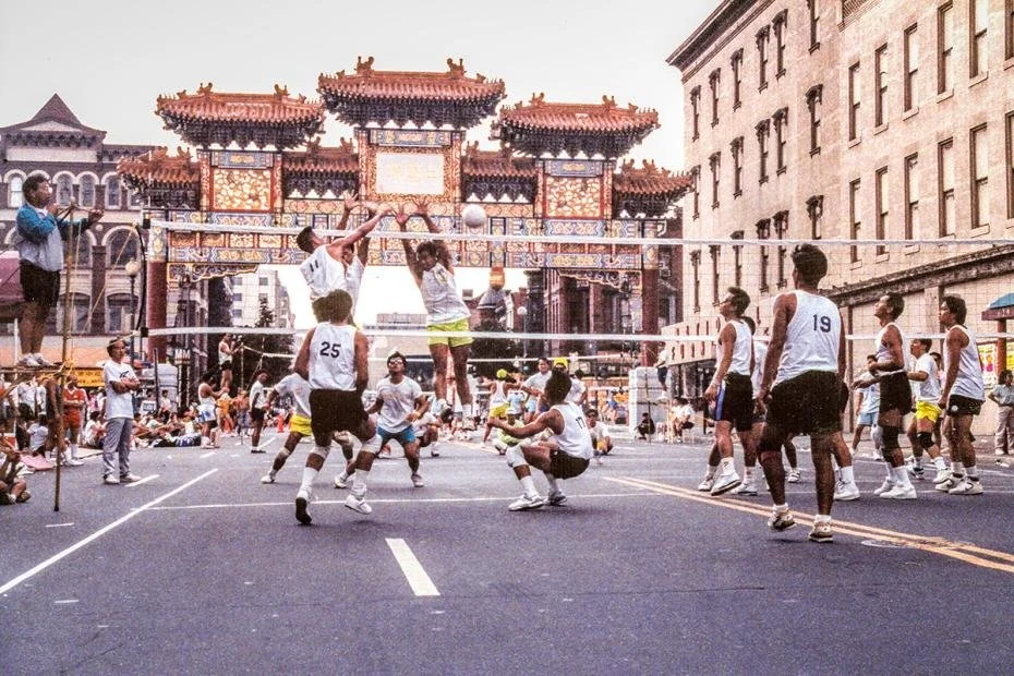 People playing volleyball on a city street during a festival with a traditional Chinese gate in the background.