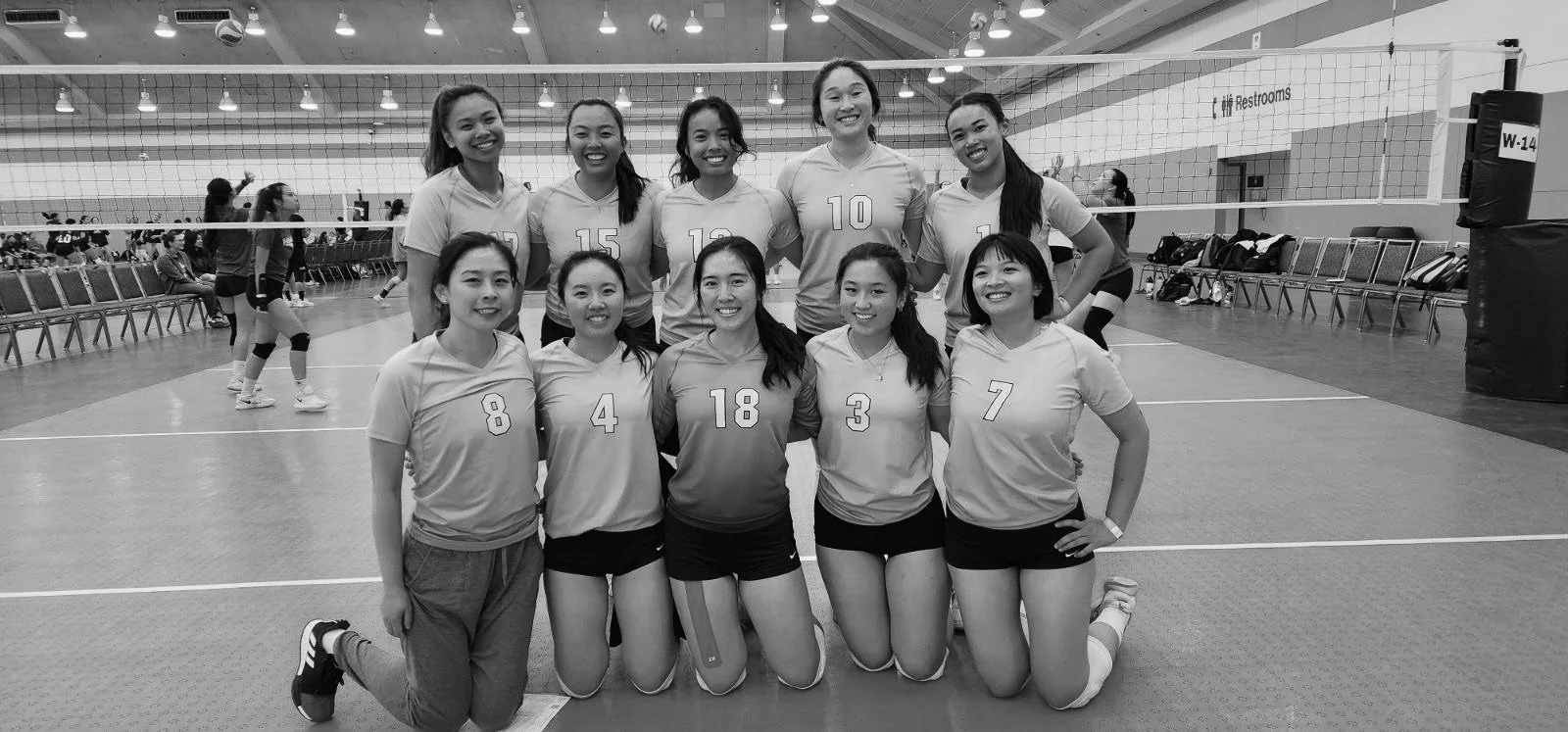 A group of 11 young women volleyball players posing together in a gymnasium, with most wearing matching jerseys with numbers. They are smiling and arranged in two rows, with some kneeling and some standing, in front of a volleyball net.
