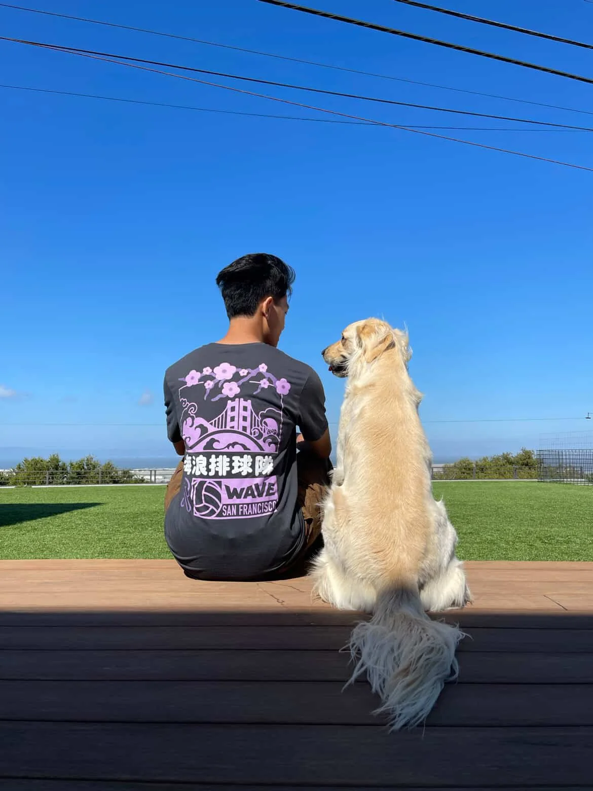 A young man and a golden retriever dog sitting together on a wooden bench outdoors, facing the blue sky with some clouds and power lines overhead, with a grassy field and ocean in the background.
