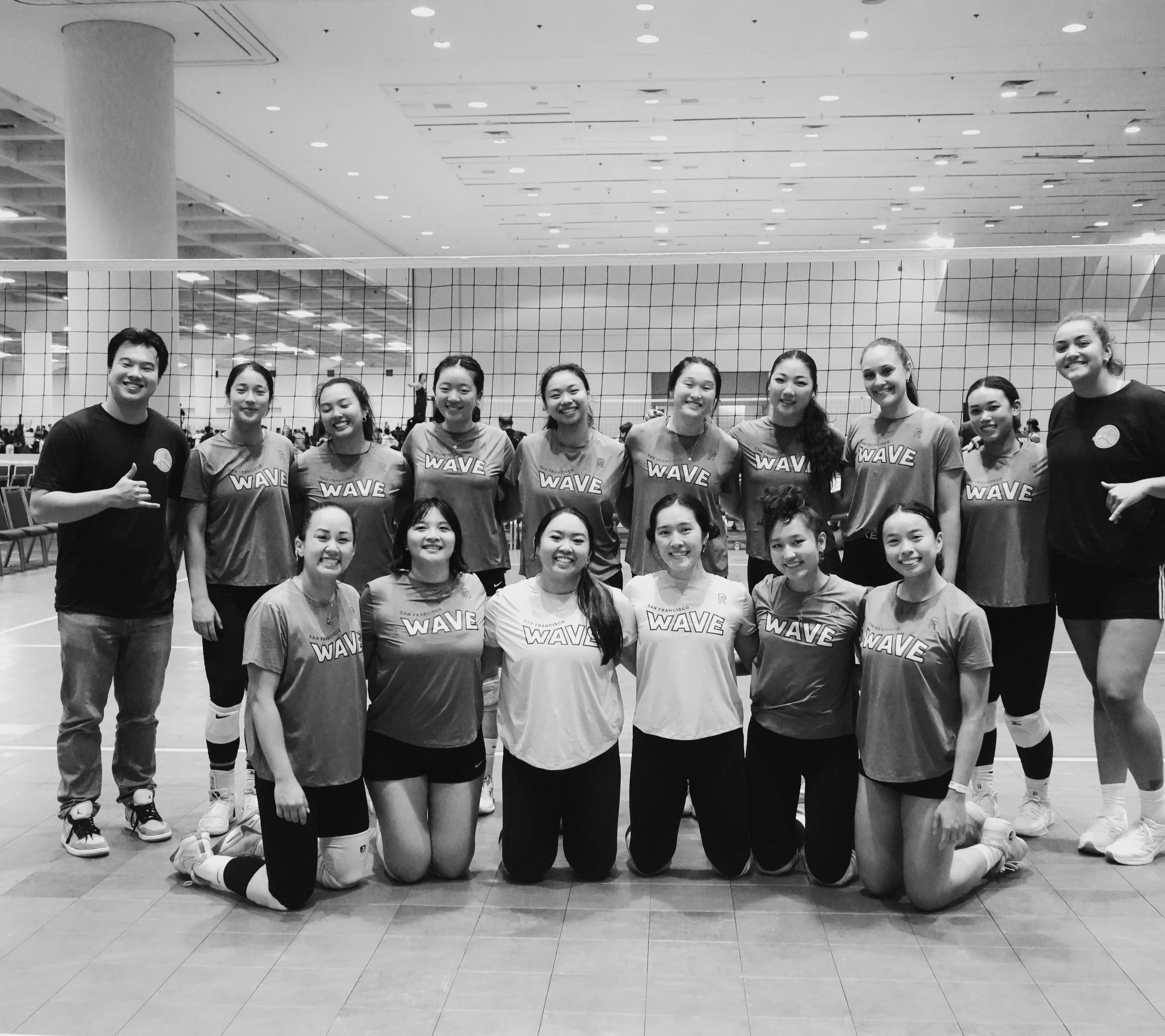 Black and white photo of a group of young women and two men, likely a volleyball team, kneeling and standing in an indoor gymnasium. The women are wearing sports jerseys with 'WAVE' printed on them, and some are wearing volleyball knee pads. The background shows the gym's ceiling with lights and a volleyball net.