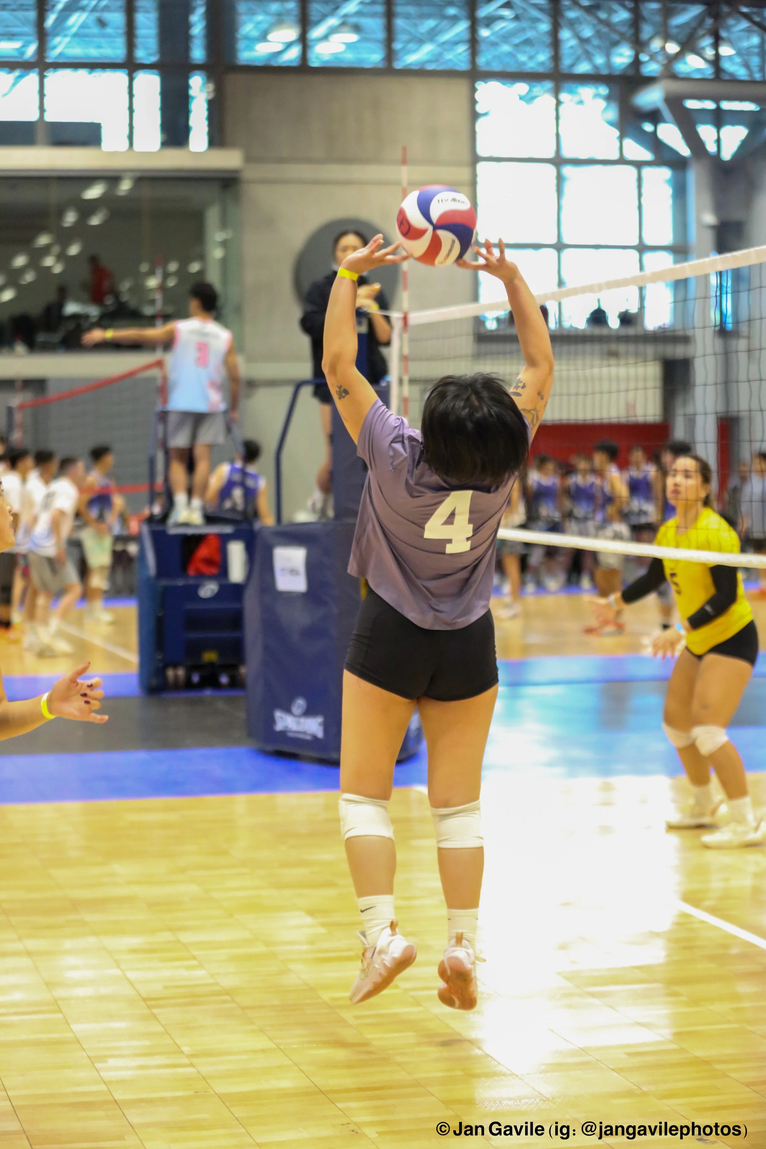 A volleyball player with a jersey numbered 4 jumping to hit a volleyball during a match in an indoor gymnasium. The photo features other players and officials visible in the background, with a high ceiling and large windows.