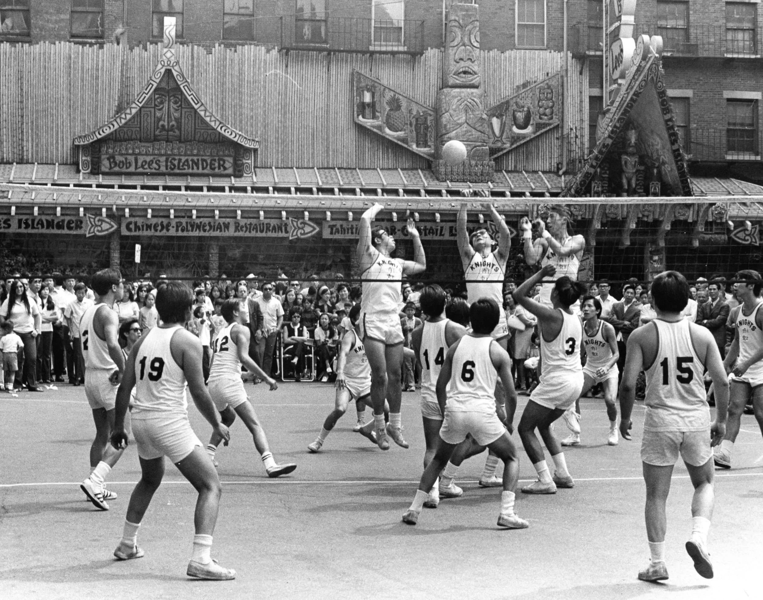A black and white photo of a basketball game with players on the court and spectators in the bleachers. There are advertising banners along the side, and four female cheerleaders or dancers in the foreground wearing matching sports uniforms.
