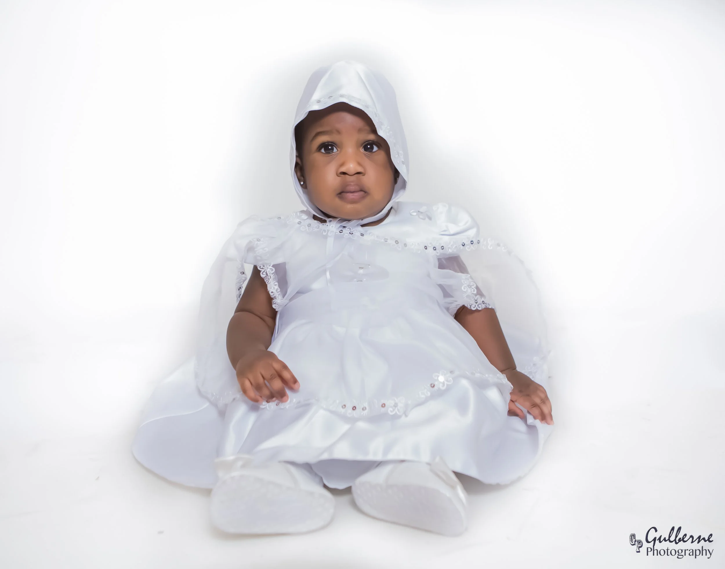 A baby girl dressed in a white dress with lace details, wearing a white bonnet, lying on a white background, looking at the camera.