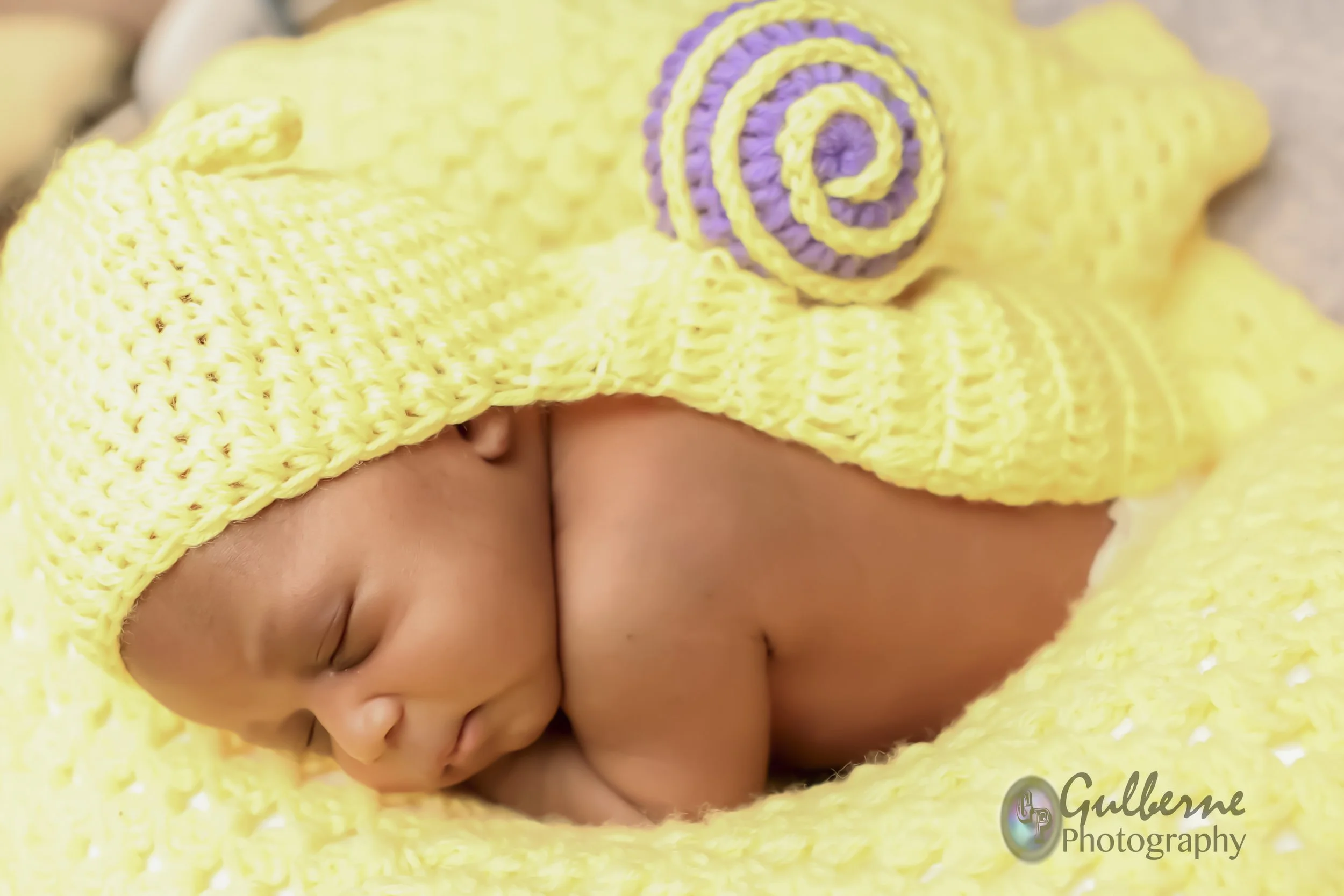 A sleeping baby wearing a yellow knitted hat with purple and yellow spiral decorations, wrapped in a matching yellow blanket, lying on its side.