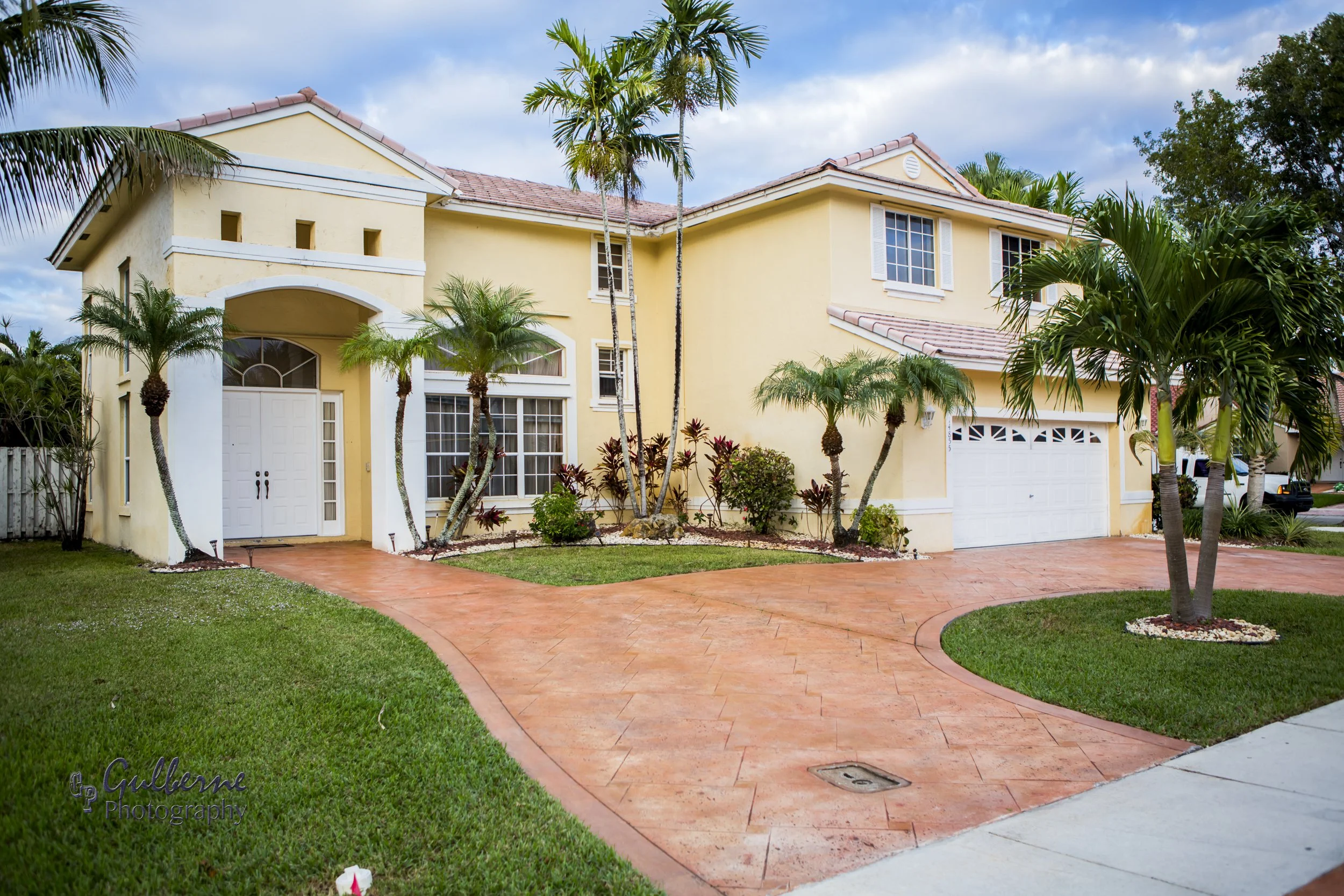 A yellow two-story house with a red tile roof, white trim, and white garage door. The house has large windows with white shutters, surrounded by palm trees and tropical plants. The driveway is made of red brick pavers, and there is a small lawn with 