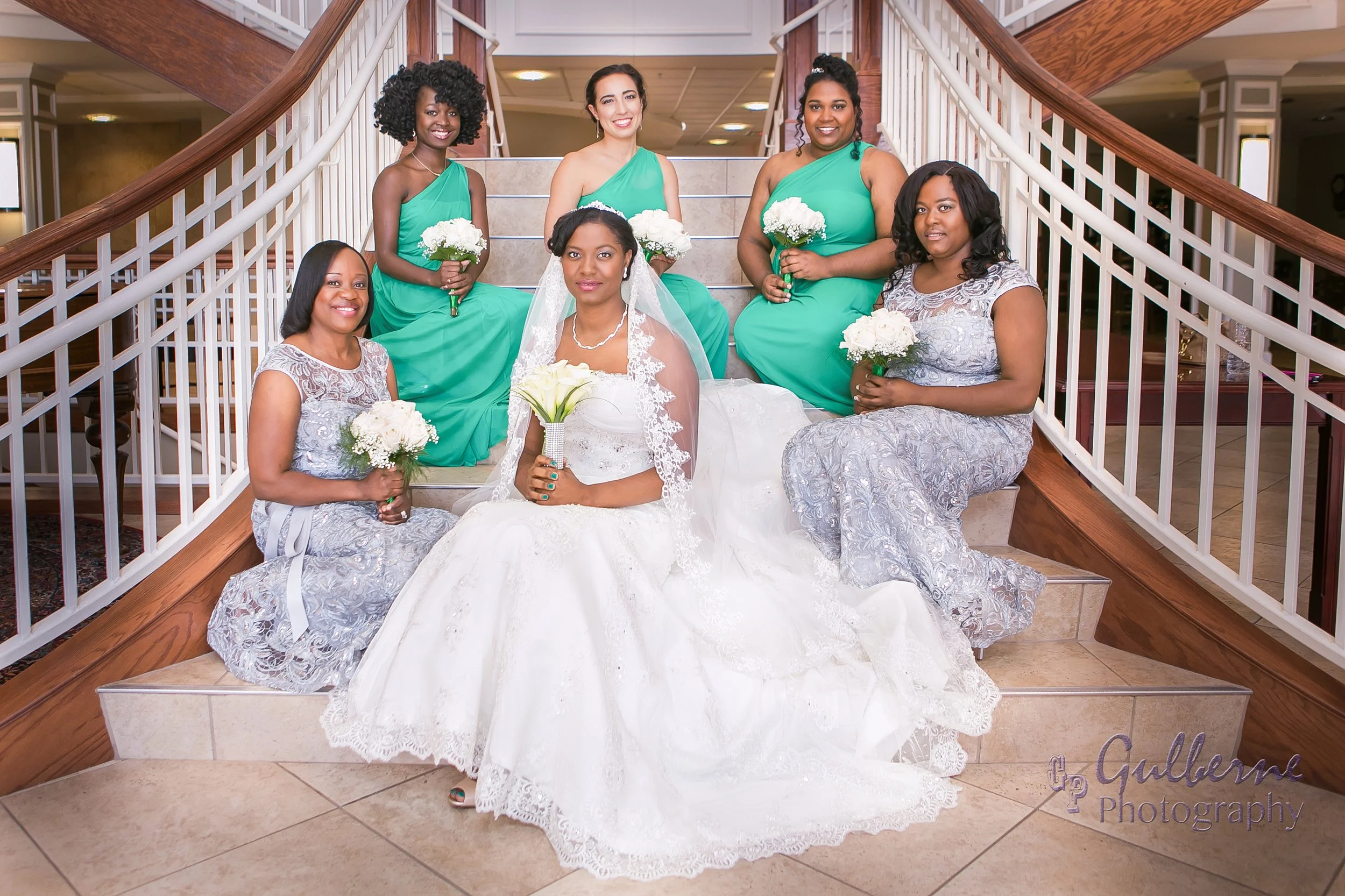 A bride in a white wedding dress holding a bouquet of white calla lilies, sitting on stairs with five women around her, four in matching green dresses holding white flower bouquets, and one in a silver lace dress, all smiling at the camera.