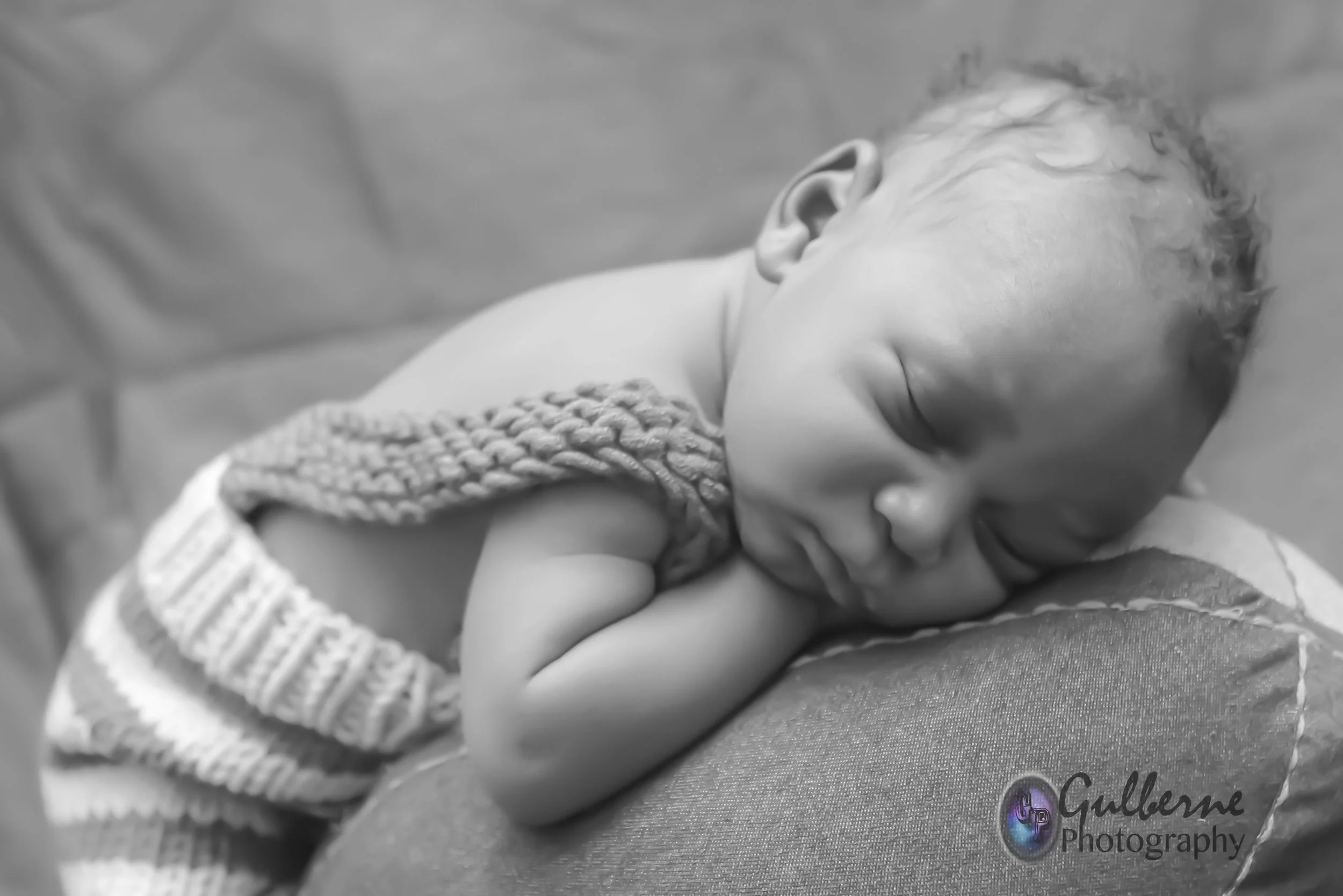 Close-up of a sleeping newborn baby with curly hair, lying on a blanket, and wearing a knitted garment, captured in black and white.