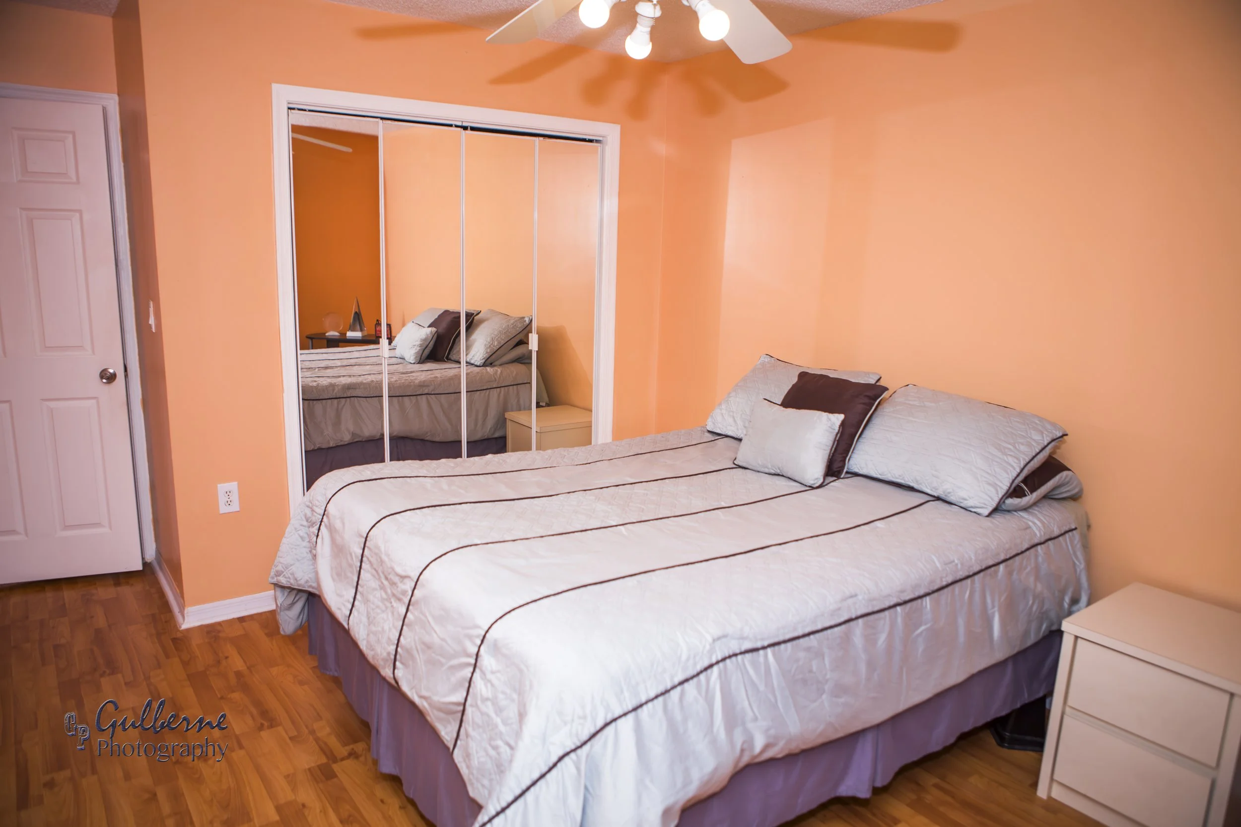 A bedroom with peach-colored walls, a bed with striped bedding and pillows, a nightstand, large mirrored closet doors, a ceiling fan with lights, and hardwood flooring.
