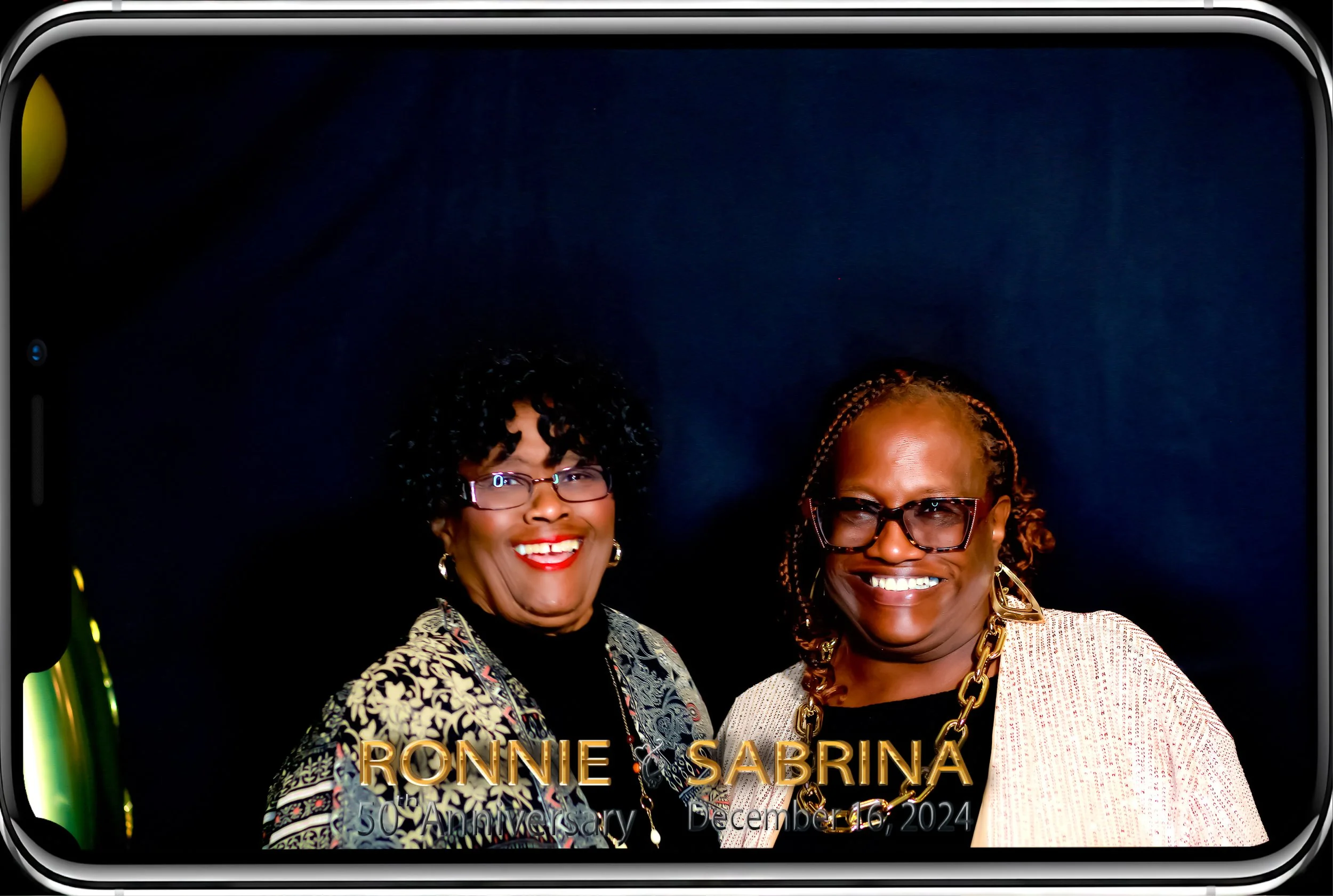 Two women smiling, one with curly black hair wearing glasses and a patterned jacket, the other with braided hair wearing glasses and a white blazer, against a dark background. The image is part of a celebration poster for the anniversary of Bonnie Sa