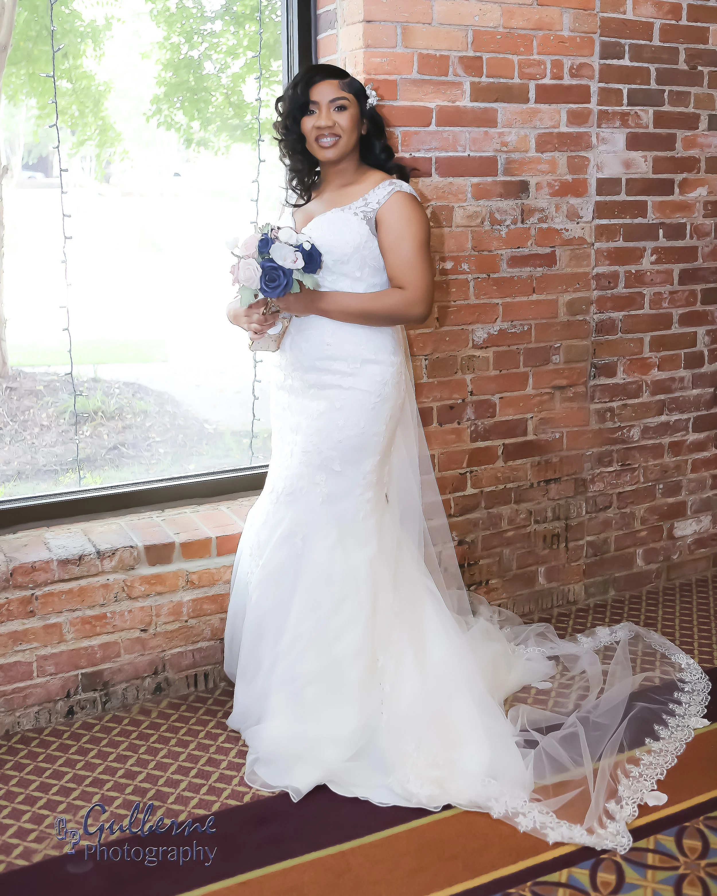 A woman in a white wedding dress holding a bouquet of blue and white flowers, standing next to a brick wall near a large window with trees visible outside.