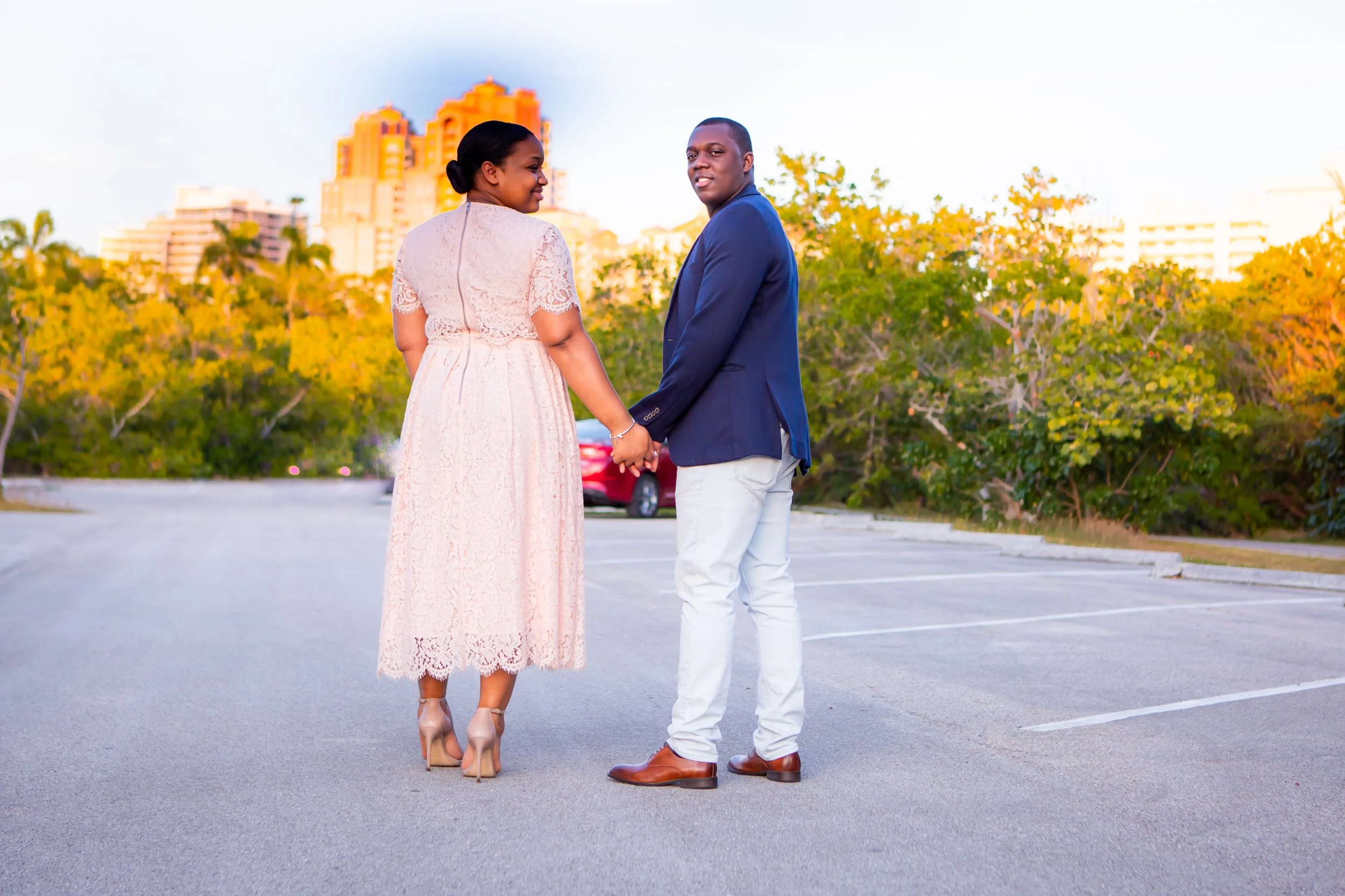 A couple holding hands in an open parking lot with trees and high-rise buildings in the background during sunset.