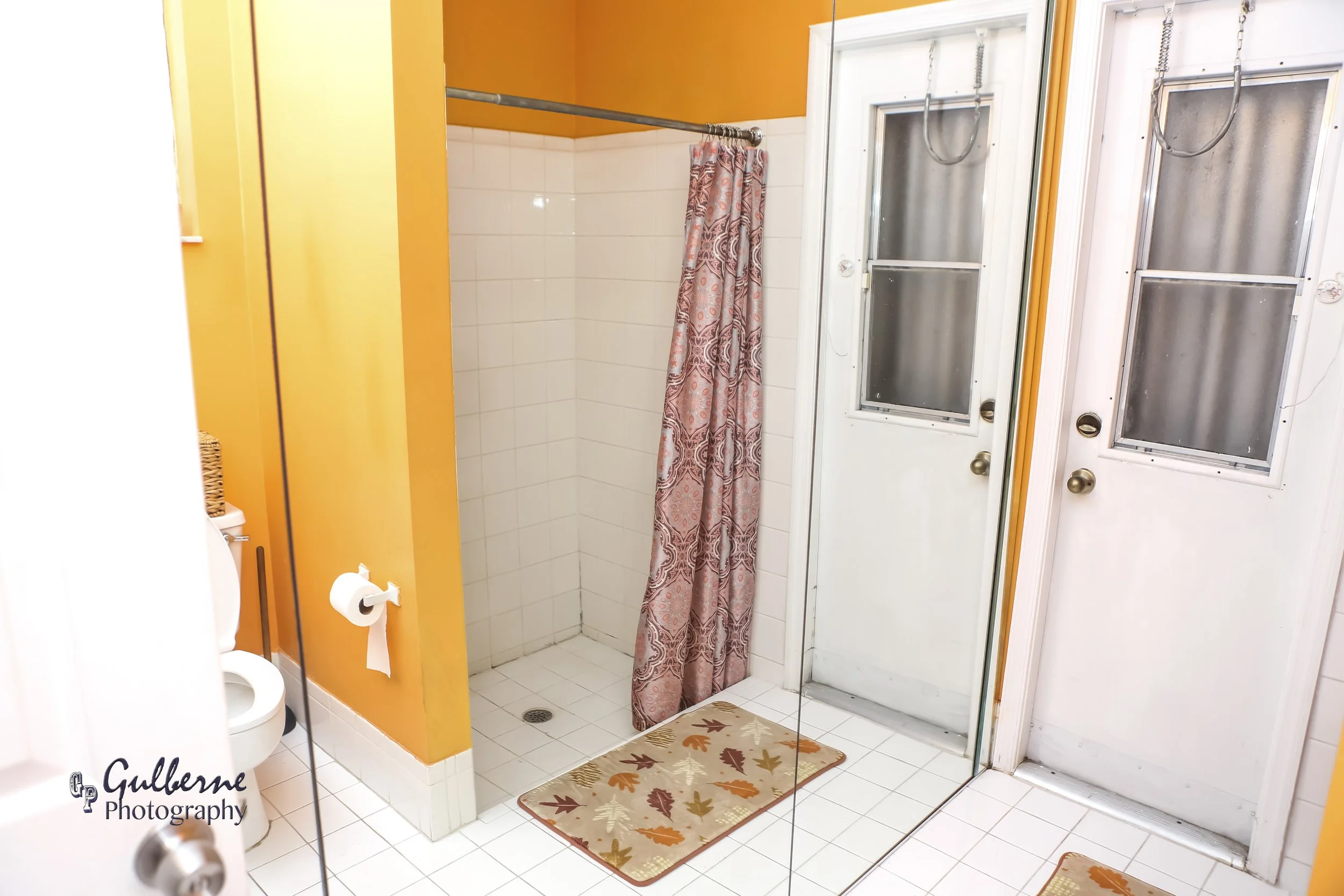 A bathroom with a white shower door, a shower curtain with a paisley pattern, and orange walls. There are two mats with autumn leaf designs on the white tiled floor, and a window with frosted glass.
