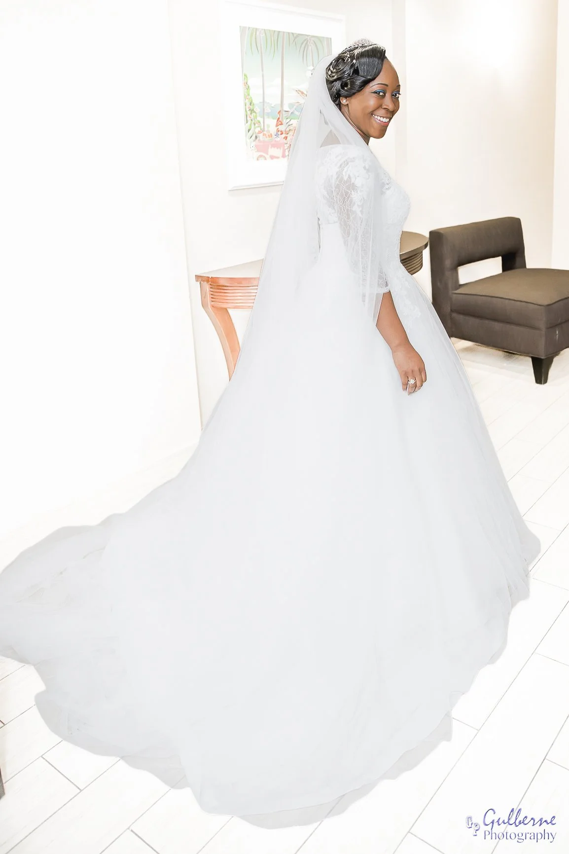Smiling woman in a wedding dress and veil standing in a bright room with white walls, a brown chair, and colorful artwork on the wall.