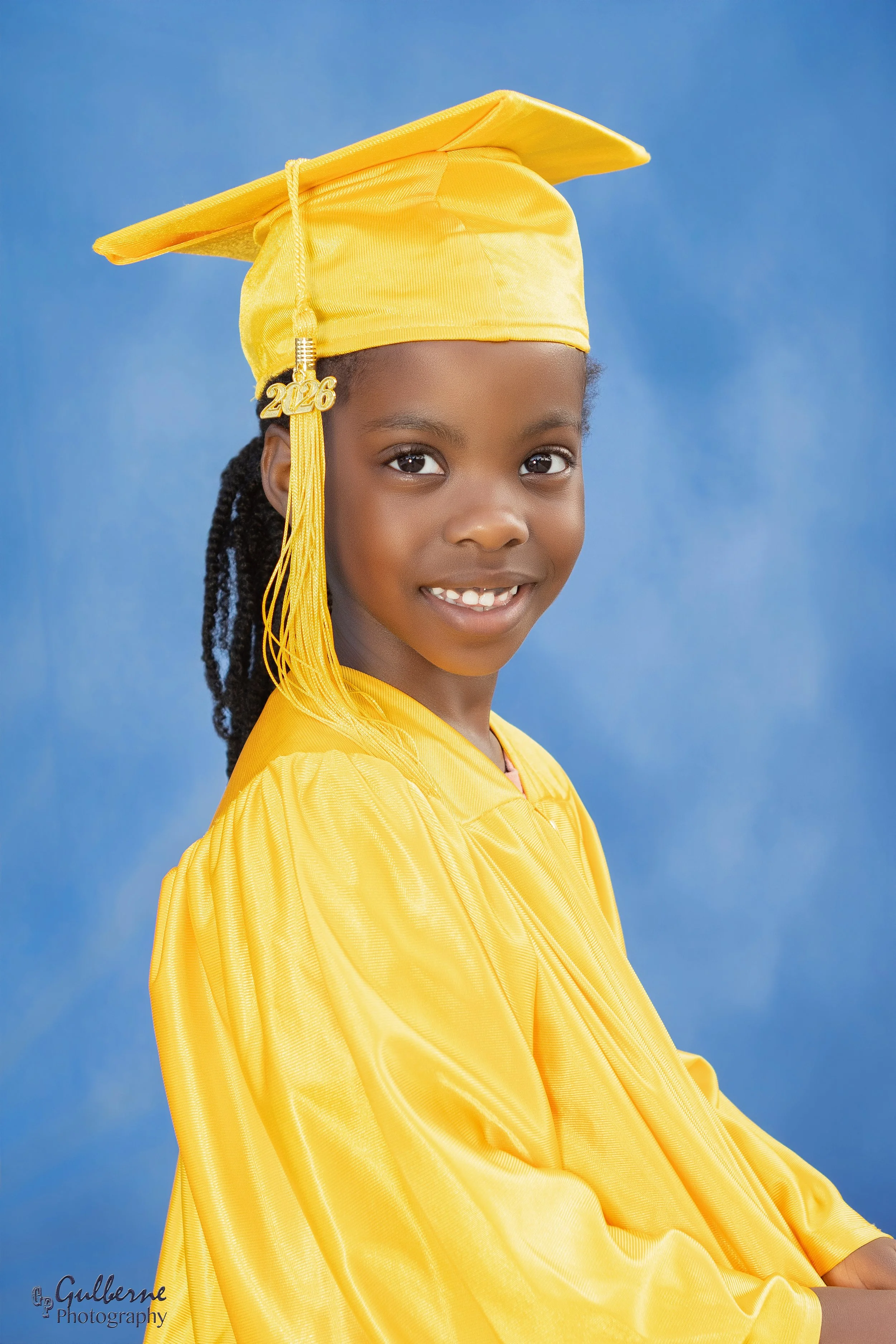 A young girl wearing a yellow graduation cap, with a gold charm that reads '2026', and a yellow gown, standing against a blue background, smiling at the camera.