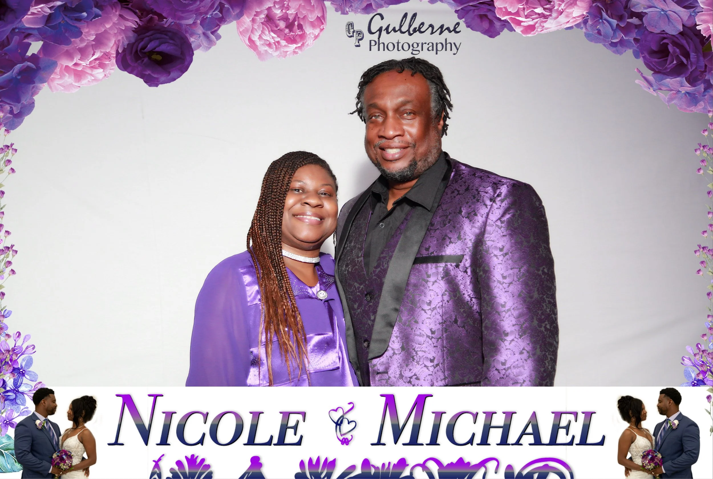 A couple in formal attire, standing under purple and pink floral decorations. The man is wearing a purple patterned suit jacket, and the woman is wearing a purple dress. The image appears to be from a wedding or engagement event.