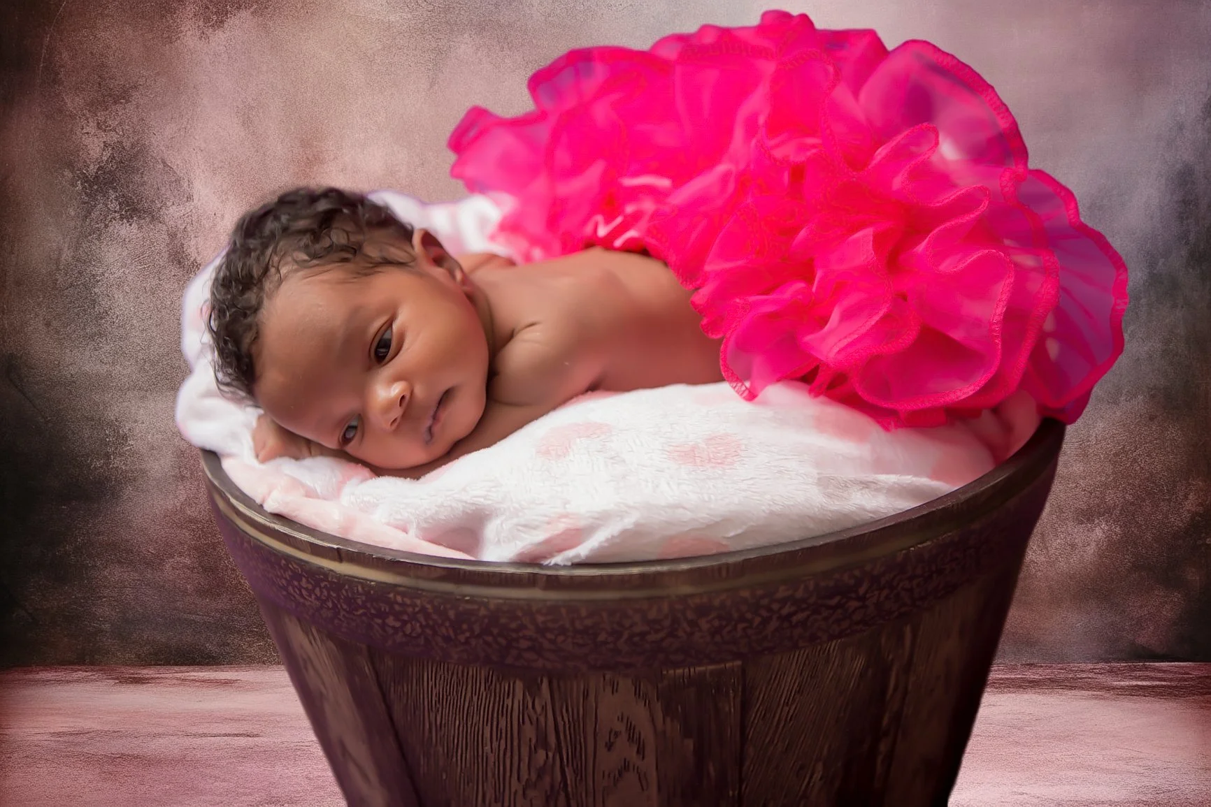 A newborn baby with dark curly hair lying on a soft blanket inside a decorative wooden bowl, wearing a bright pink ruffled skirt, with a beige background.