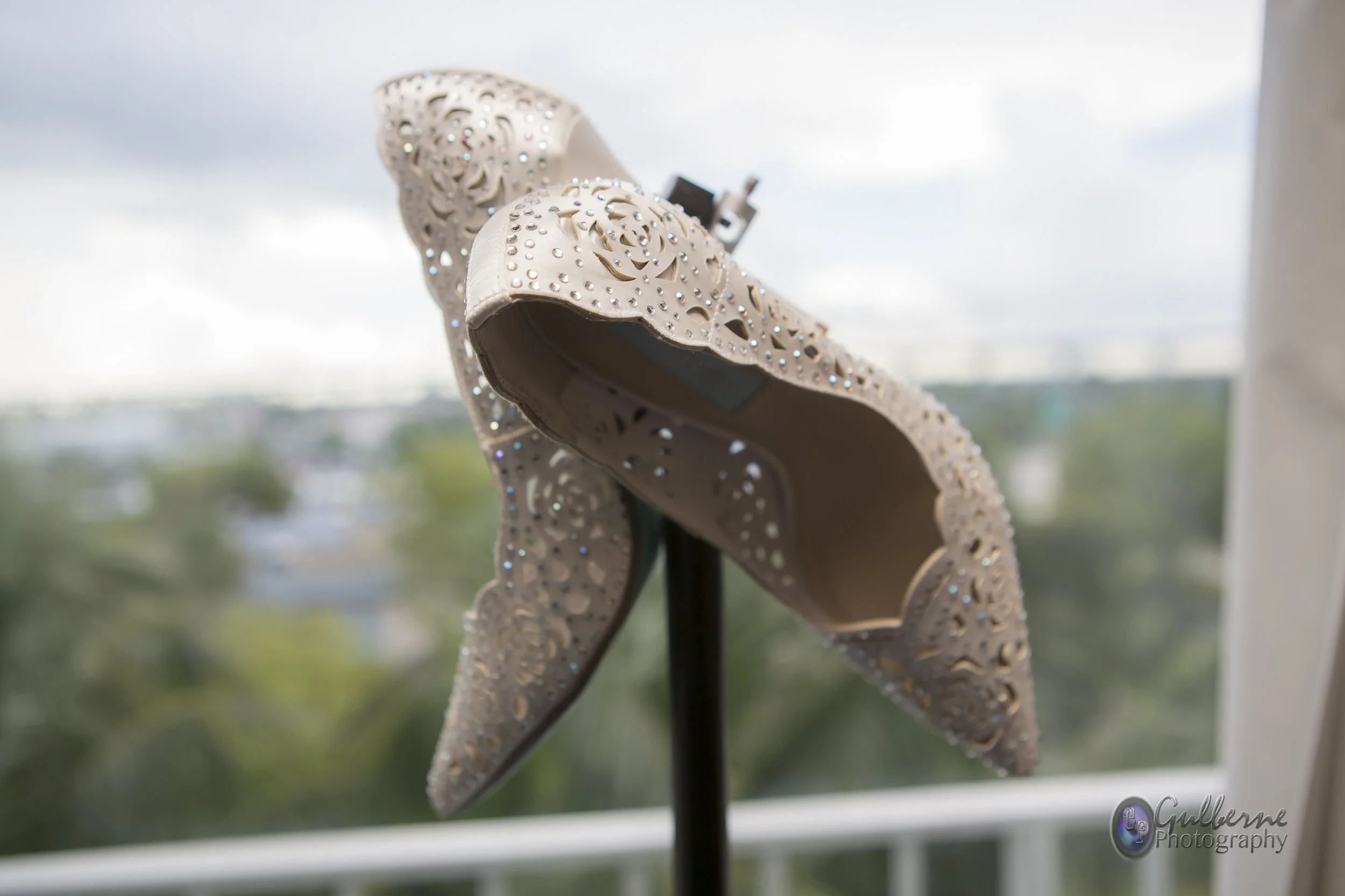 A close-up of a decorative, beige, high-heeled shoe with intricate laser-cut floral patterns and embedded rhinestones, displayed indoors near a window.