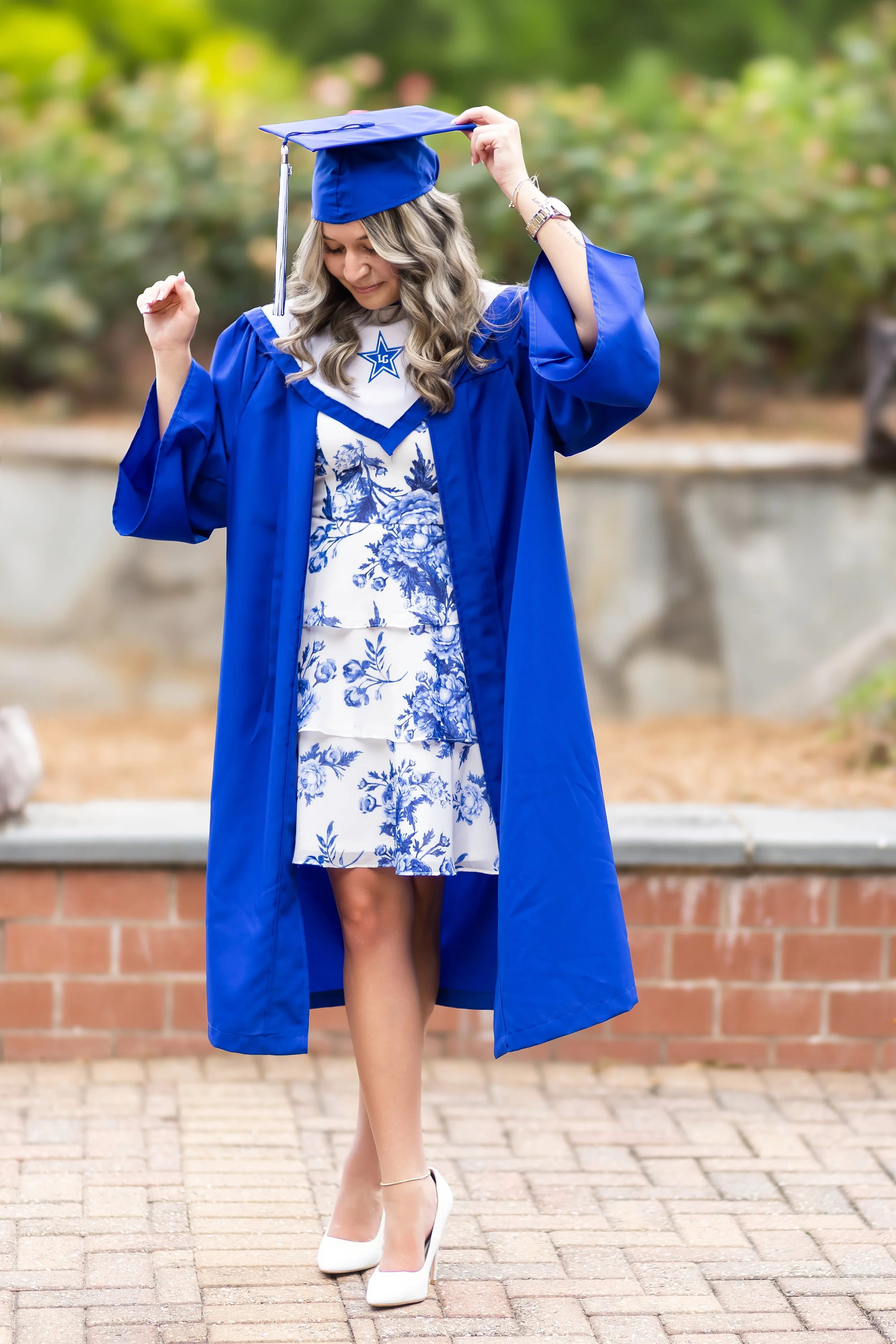 Young woman in a blue graduation gown and cap, standing outdoors on a brick path, adjusting her cap with a background of blurred greenery.