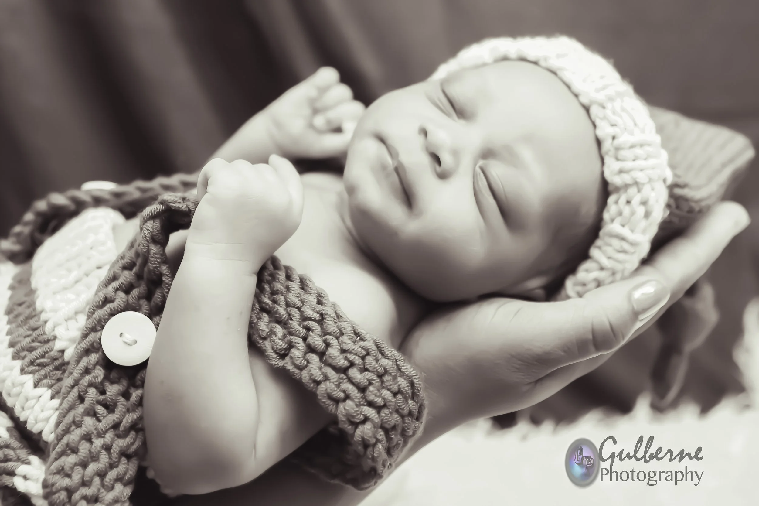 Close-up of a sleeping baby with a headband, being gently held by an adult hand, surrounded by crocheted blankets.