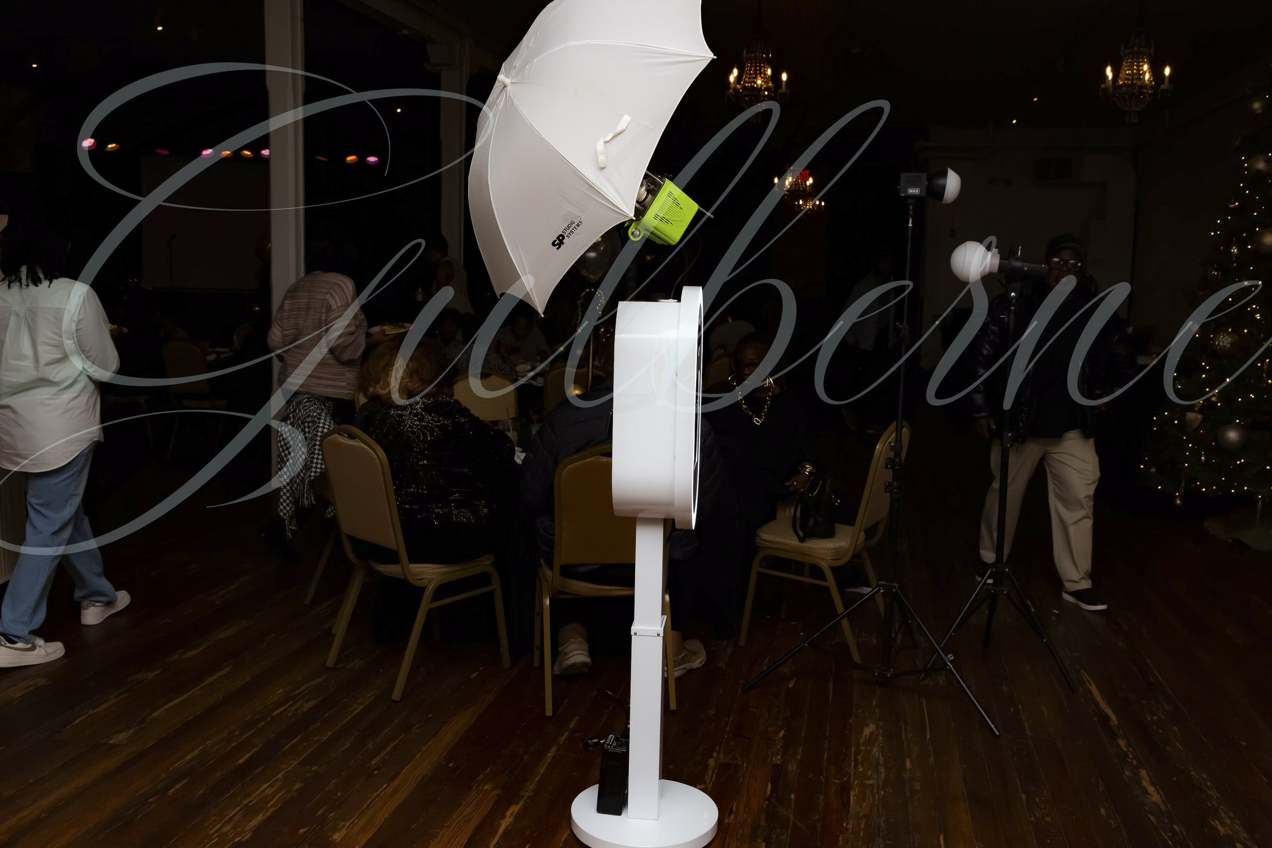 Photo of a photo booth with a white umbrella light, multiple chairs, and people in the background at an indoor event.