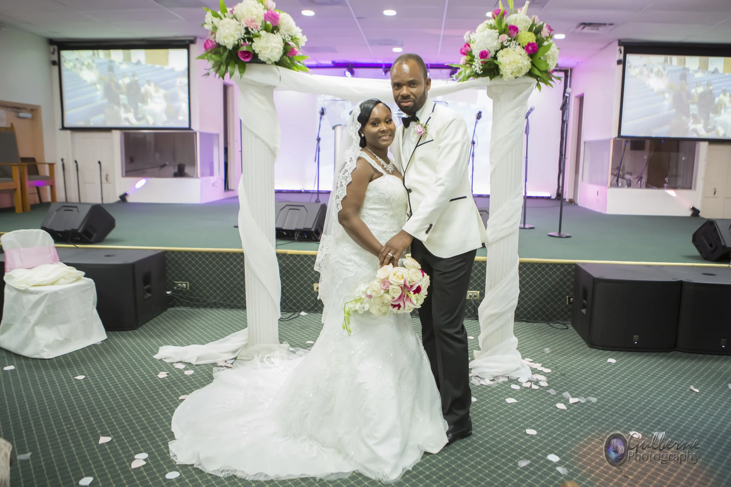 A bride and groom standing under a decorated wedding arch with flowers, holding hands and posing for a photo in a banquet hall with large screens in the background.