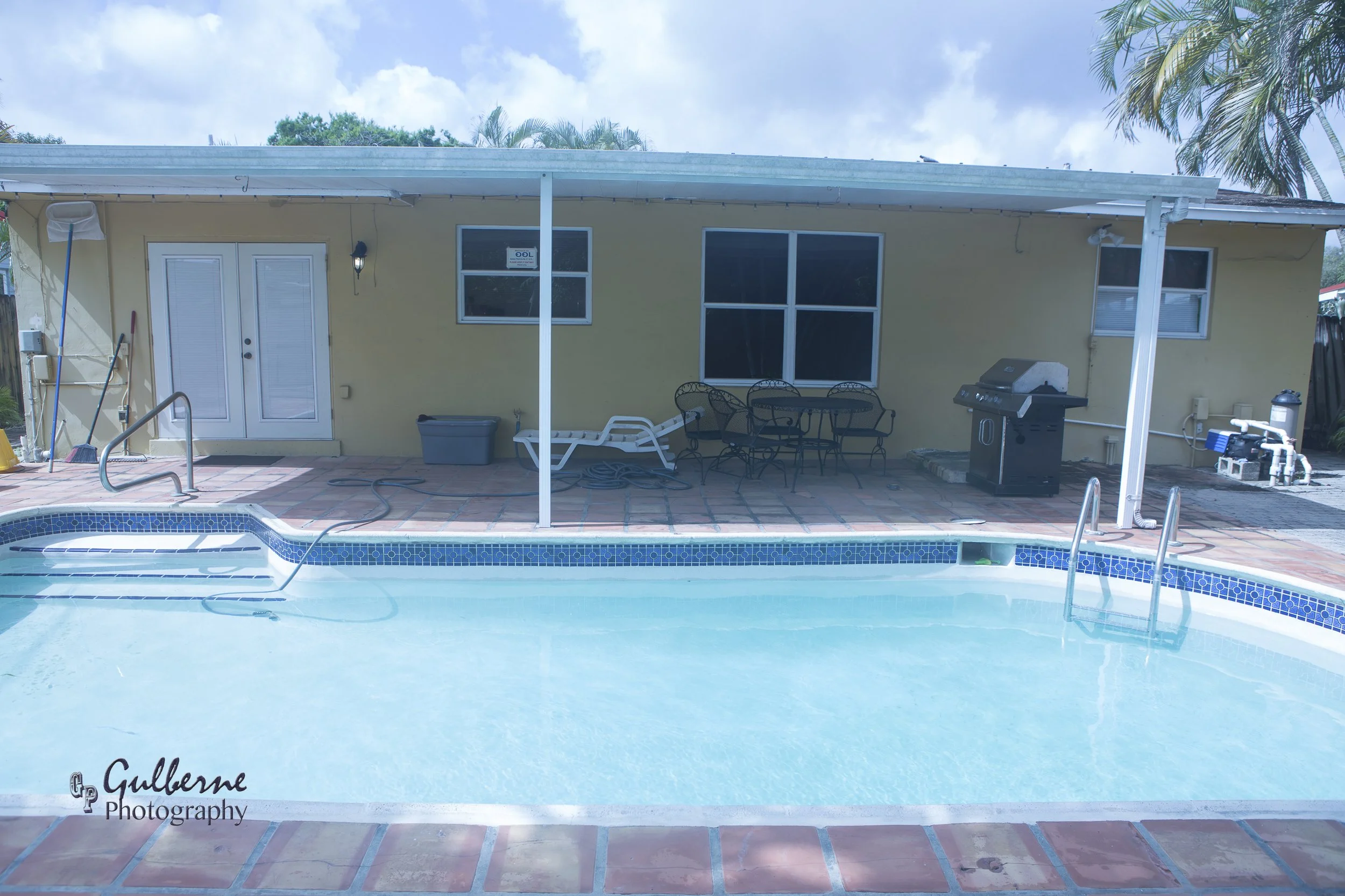 Backyard with swimming pool, patio furniture, grill, and house with yellow walls and multiple windows. Palm trees and cloudy sky in background.