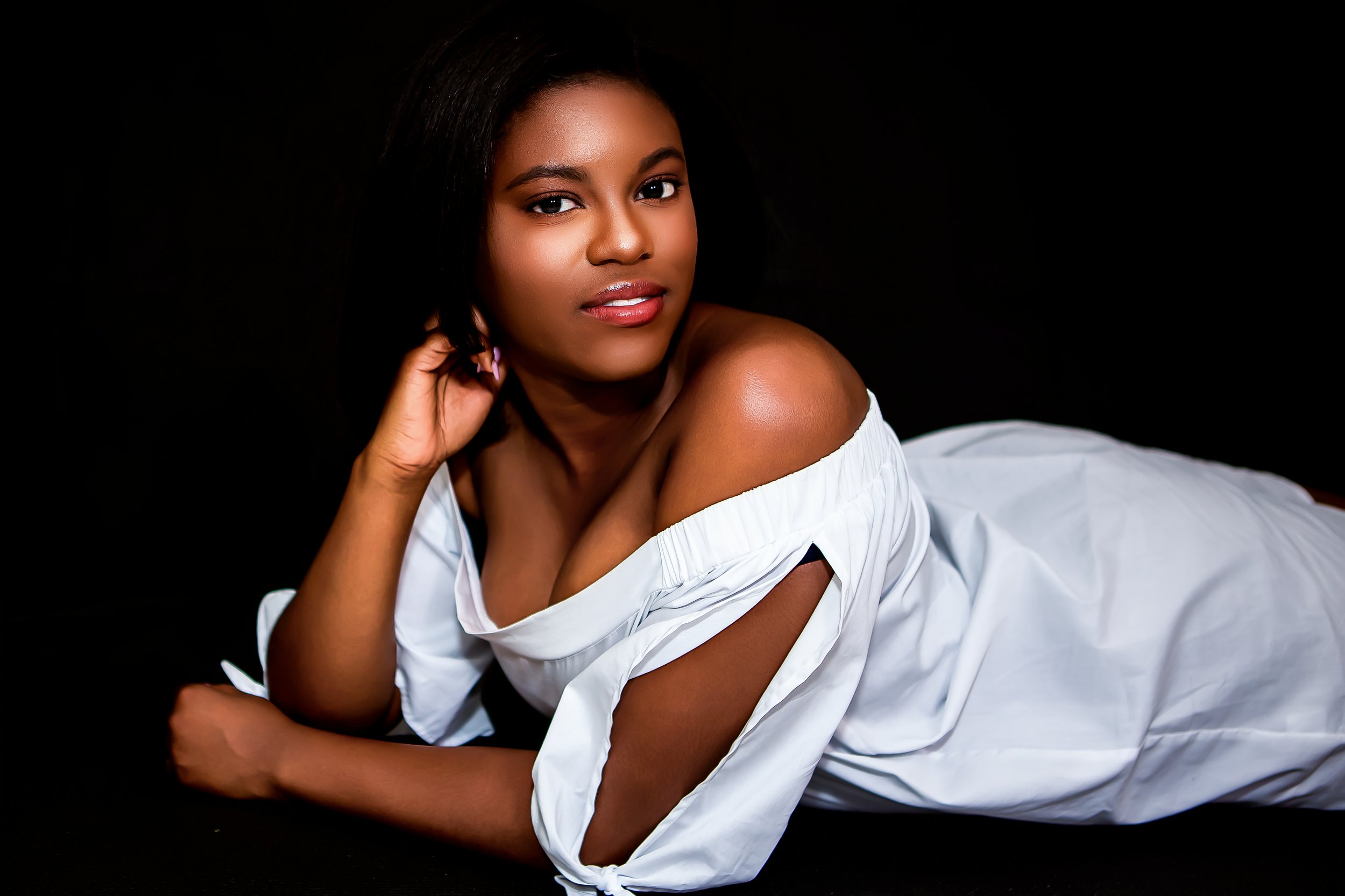 A young woman with dark hair and brown skin lying on a white surface, posing with her head resting on her hand against a black background, wearing a white off-the-shoulder top.