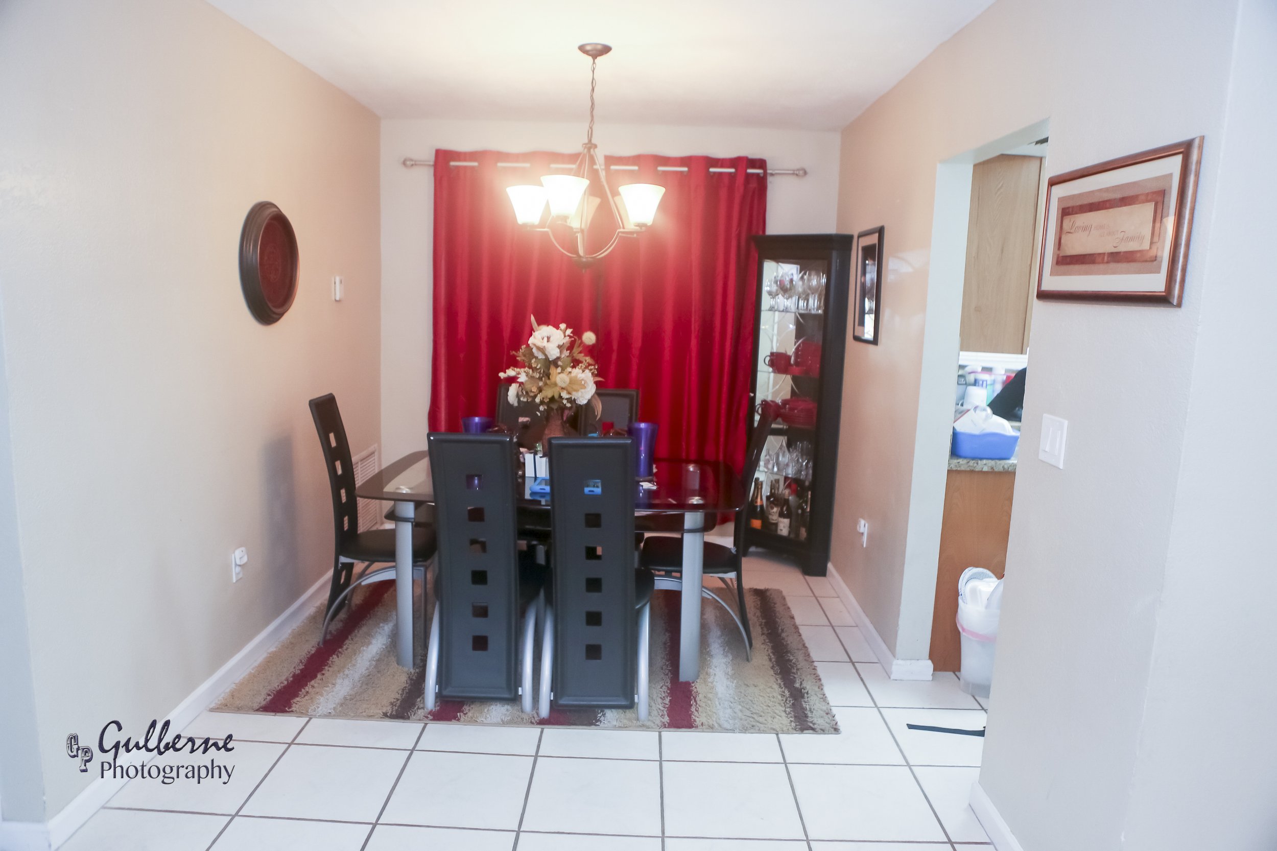 A dining room with a dark wood table and six black chairs. Red curtains cover a window behind the table. A chandelier with four lights hangs above the table. A tall glass cabinet with glassware is in the corner. A vase with white and beige flowers is