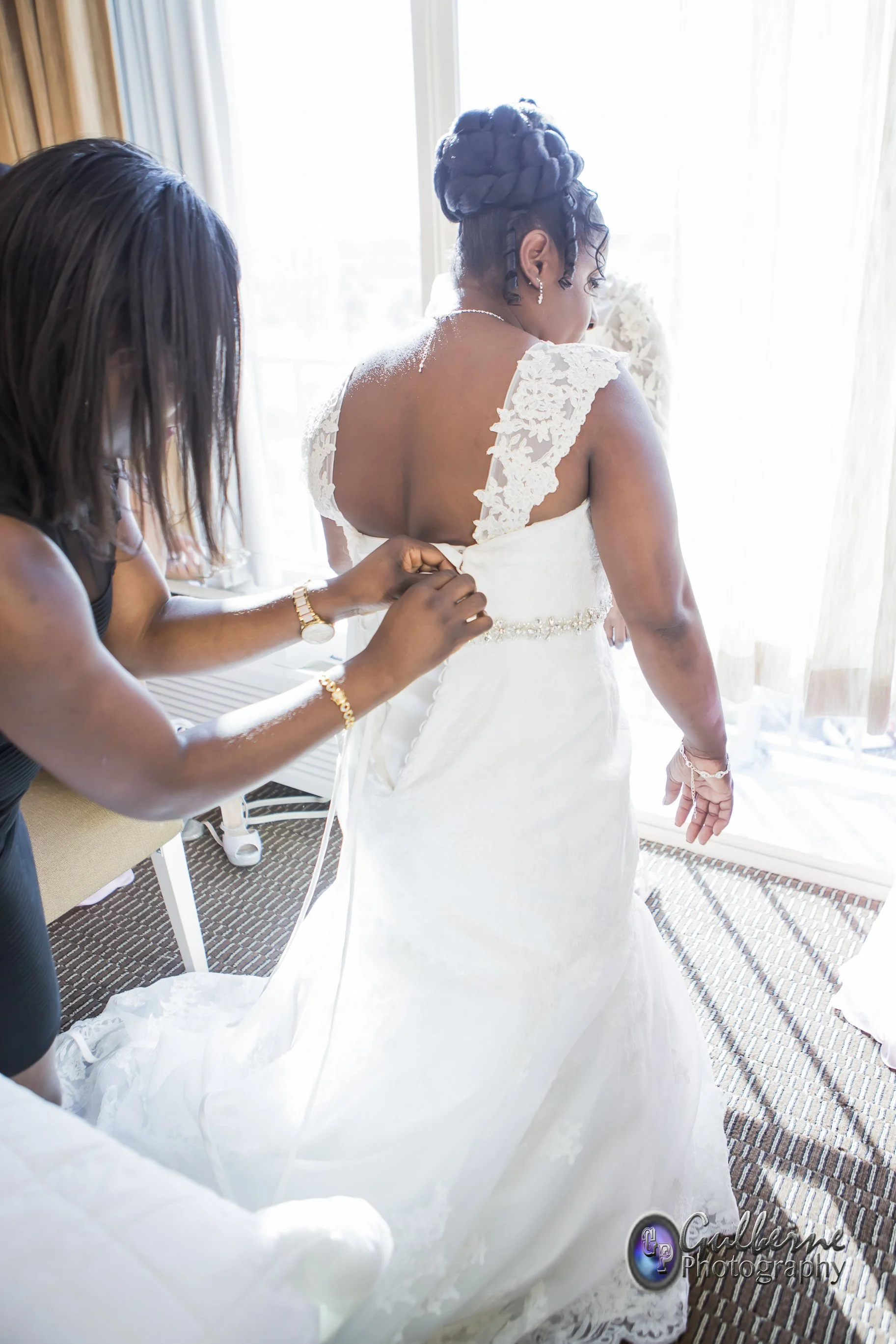 A bride in a white lace wedding dress getting dressed with help from a woman, while standing near a window with sheer curtains, in a hotel room.