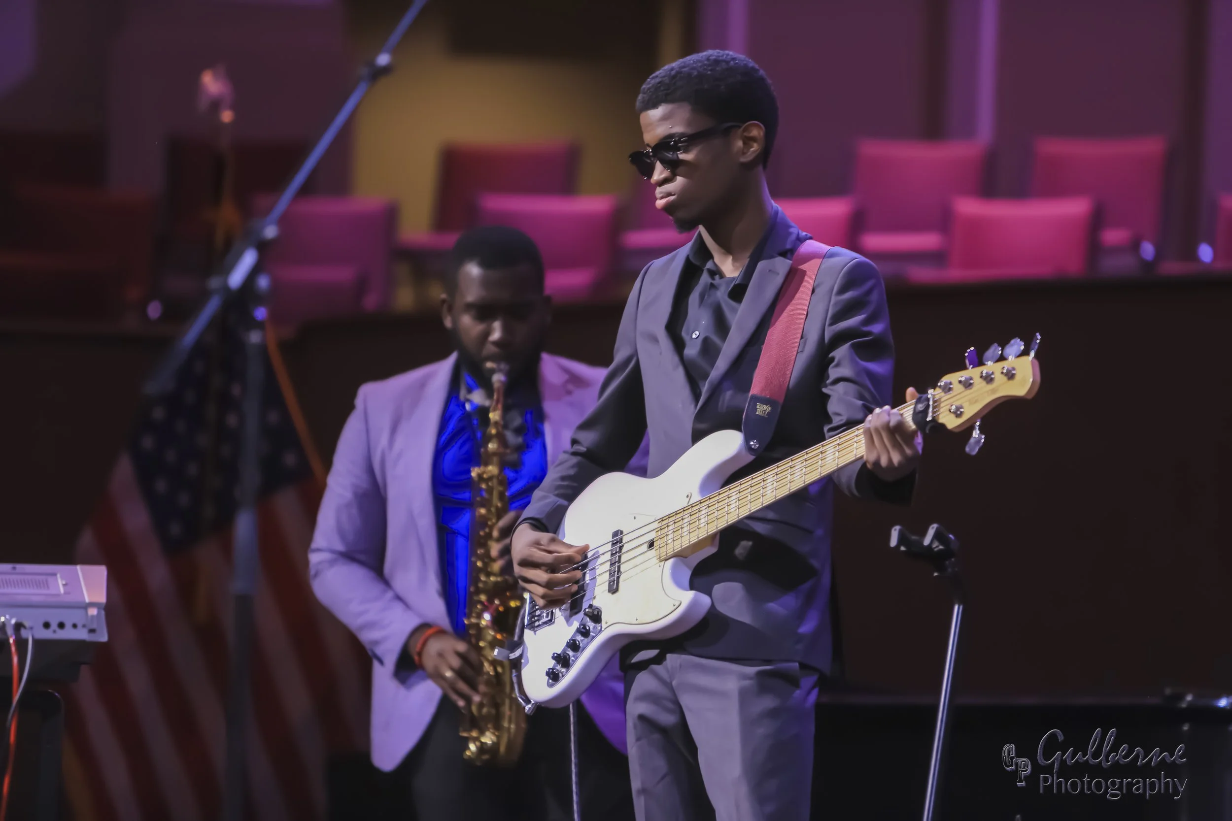 Young man wearing sunglasses playing an electric guitar on stage, with another musician playing a saxophone in the background, all in a setting with pink chairs and stage lighting.