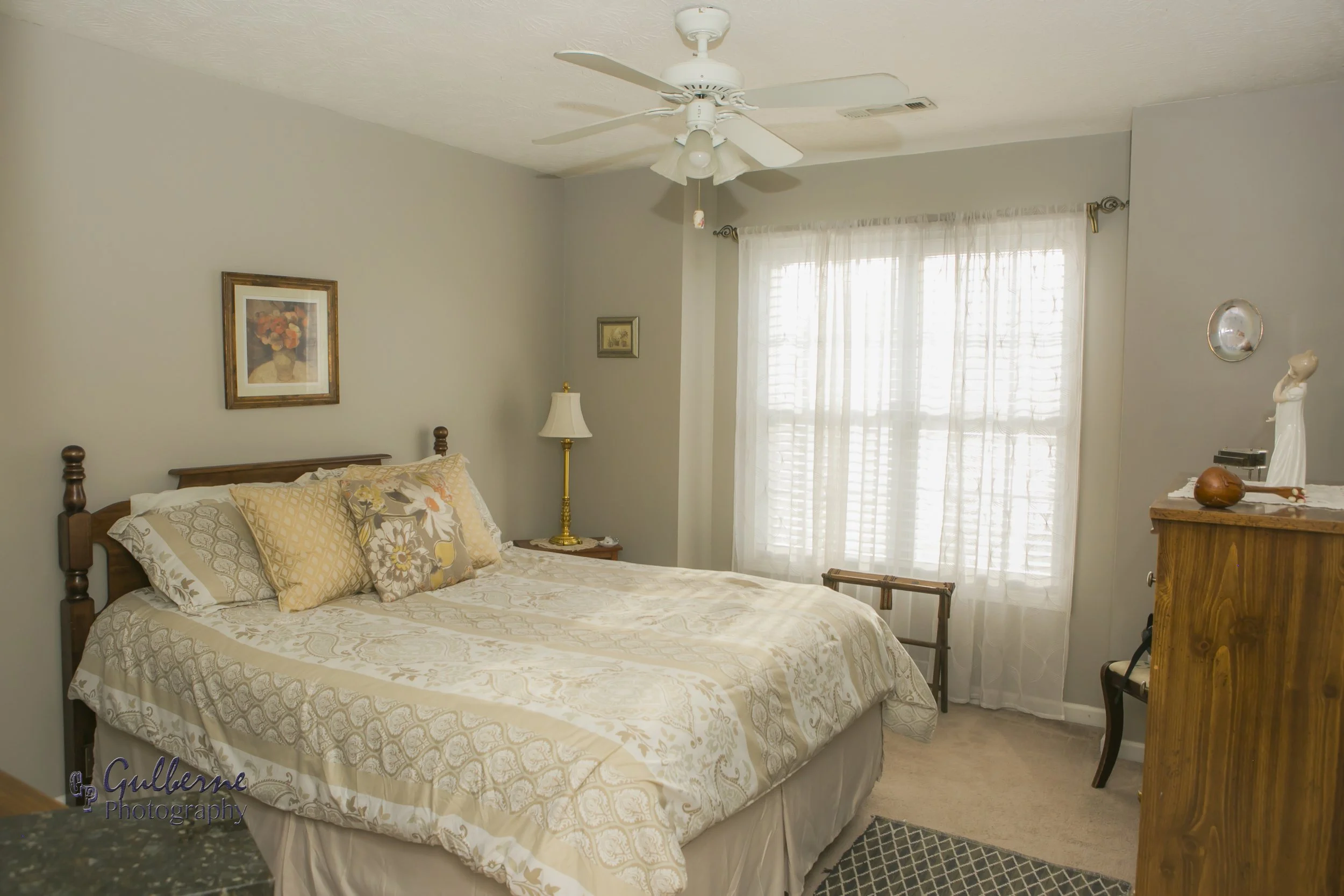 A cozy bedroom with a wooden bed frame, patterned bedding, and decorative pillows. There is a wooden dresser with a small statue on top, a gold lamp, framed pictures on the walls, sheer white curtains on the window, and a ceiling fan.