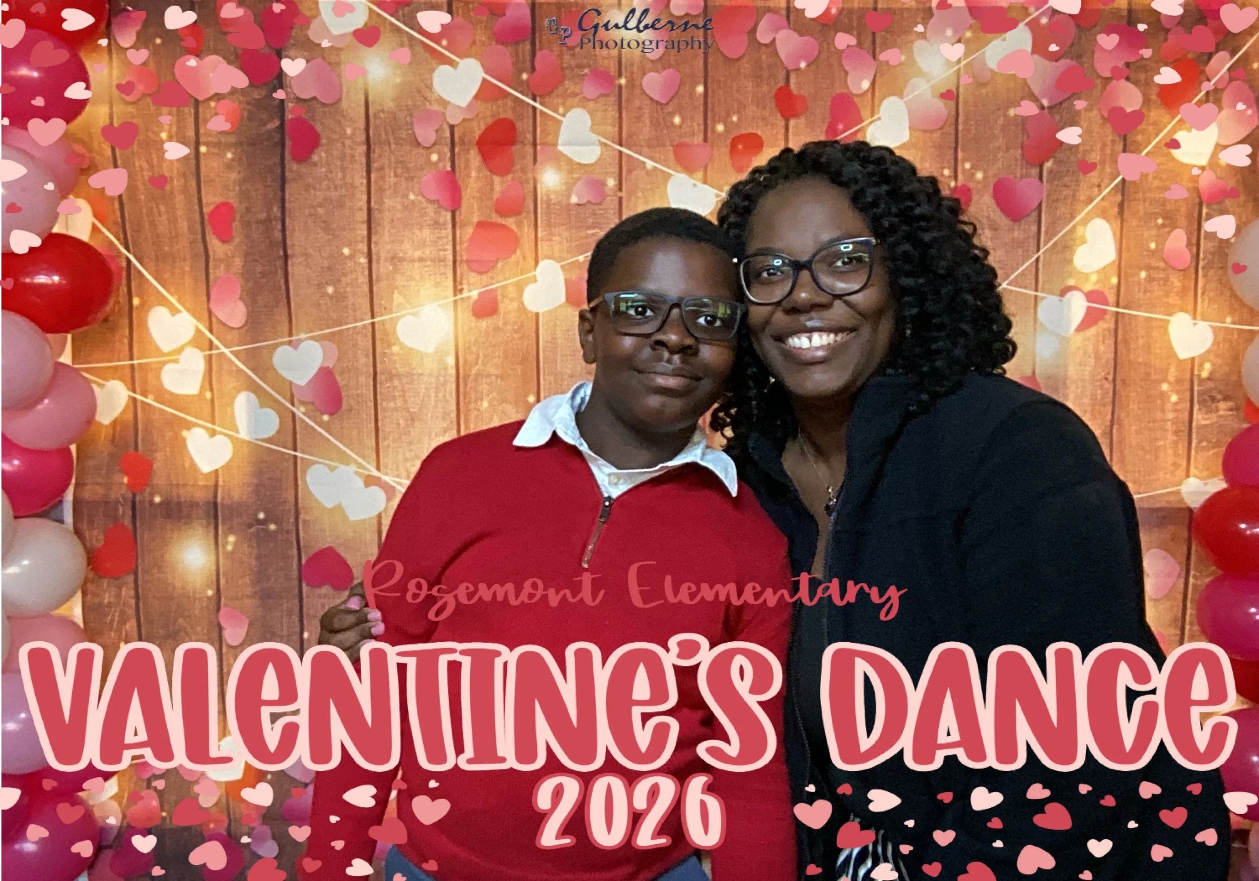 A woman and a young girl standing together at a Valentine's Day event. The background is decorated with wooden panels, pink, red, and cream balloon hearts, and string lights. The woman is smiling, wearing glasses and a black top, while the girl, also