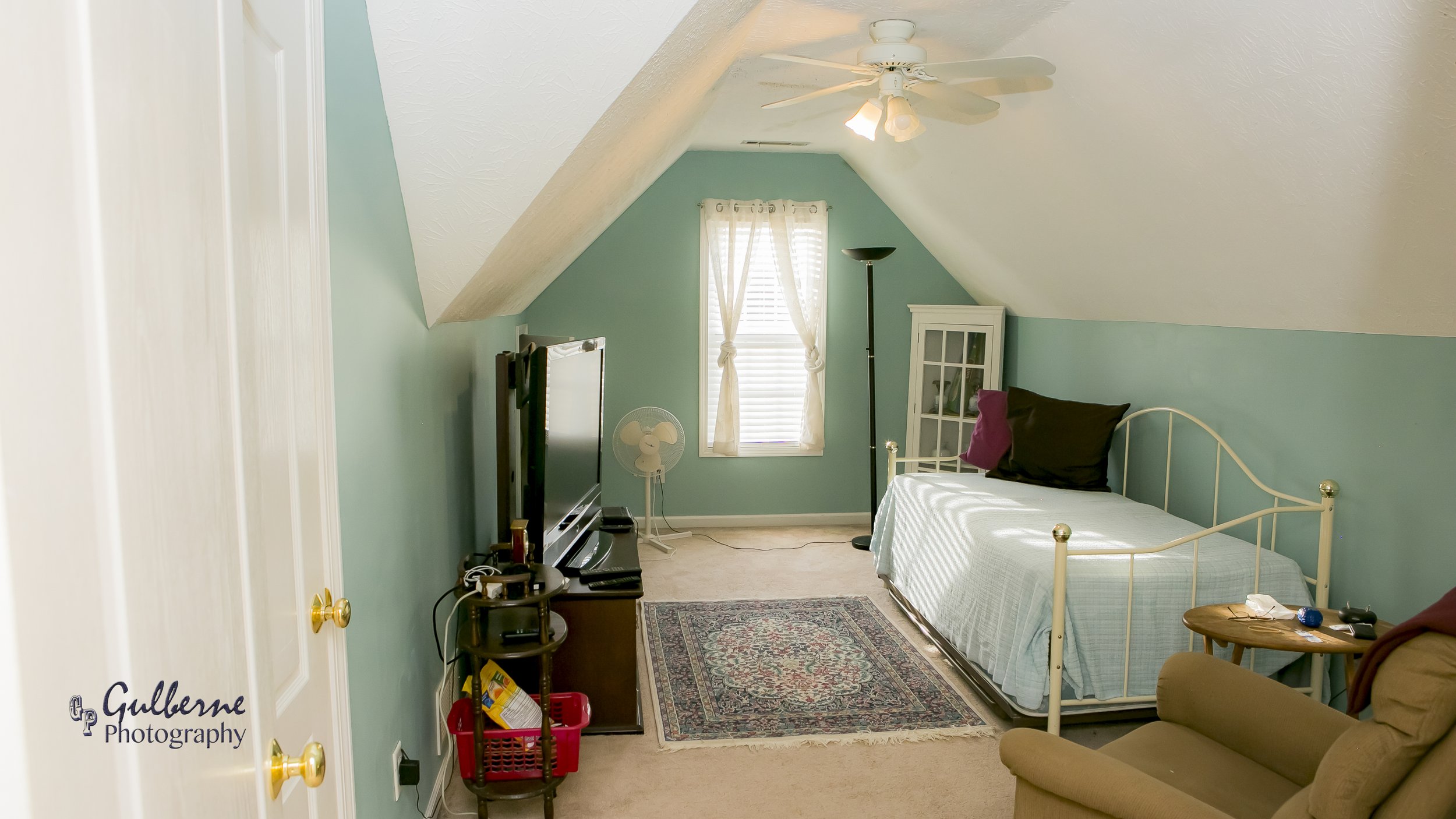 A cozy bedroom with teal walls and sloped ceiling. There's a window with sheer curtains, a bed with a metal frame and pillows, a small wooden side table, a standing lamp, a fan, and a television.