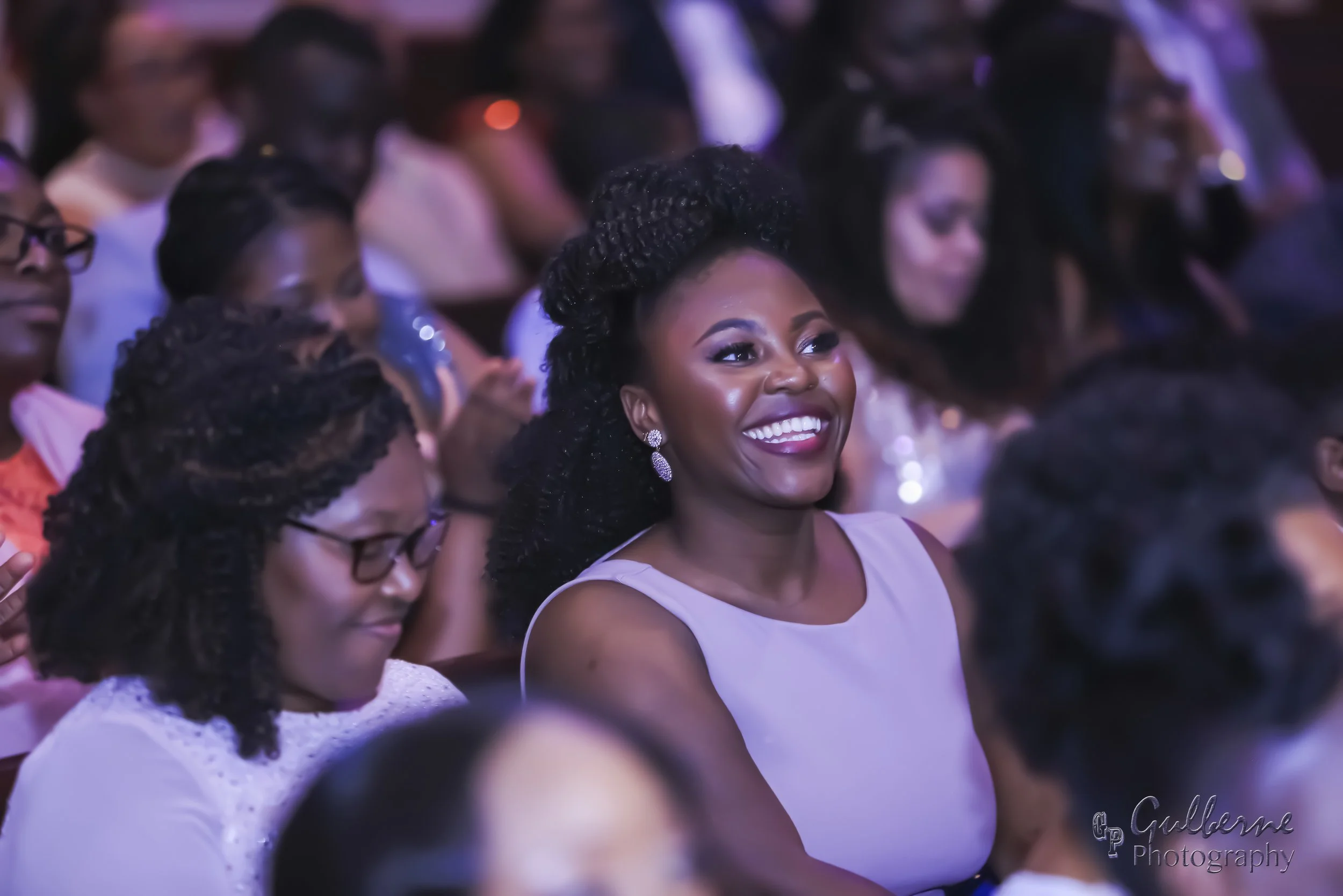A group of people attending an event, with a woman in the foreground smiling and wearing a light-colored dress and earrings, surrounded by others in the audience.