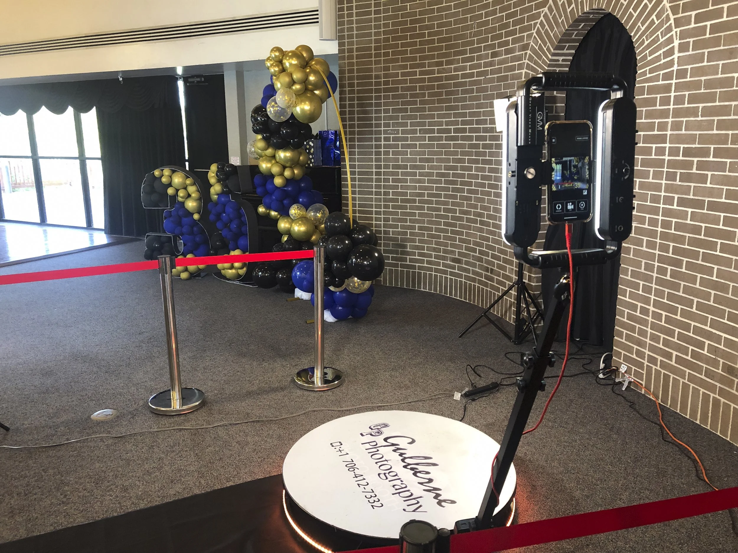 Photo booth decorated with black, gold, and blue balloons, with a red velvet rope barrier, and a tripod with a camera or scanner in front of a brick wall, at an indoor event or party.