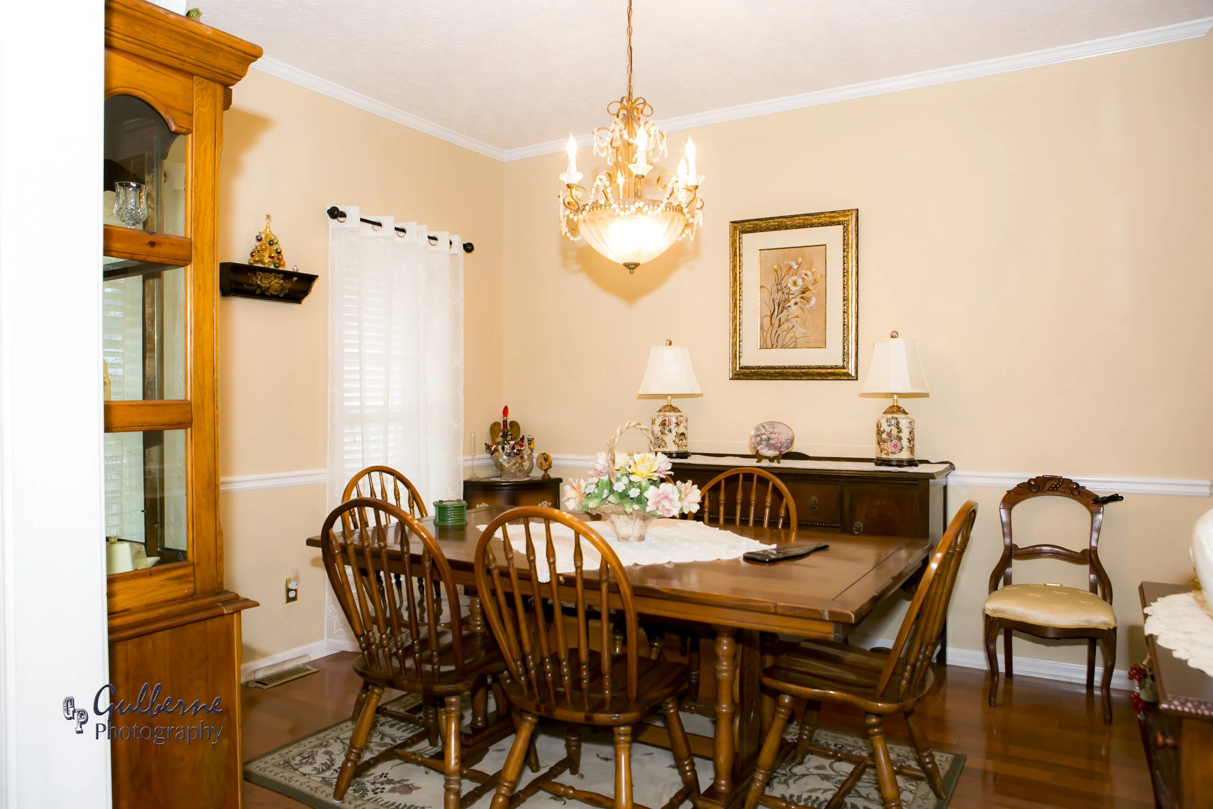 A dining room with a wooden dining table surrounded by matching chairs, a chandelier hanging from the ceiling, and a framed floral artwork on the wall. A china cabinet on the left holds glassware, and a sideboard with lamps and decorative items is ag
