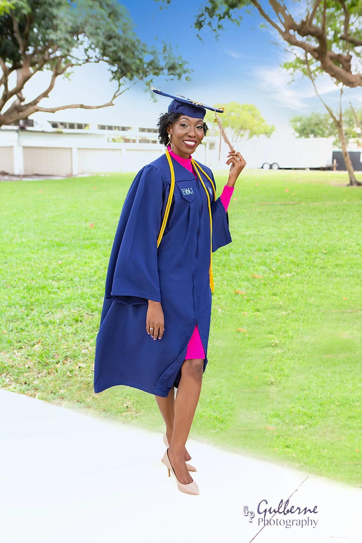 A woman dressed in a blue graduation gown and cap, with a pink dress underneath, standing outdoors on a grassy area with trees and a white fence in the background. She is smiling, holding her cap with her left hand, and wearing high heels and gold jewelry. The graduate is wearing a gold honor cord around her neck and an embroidered badge on her gown.