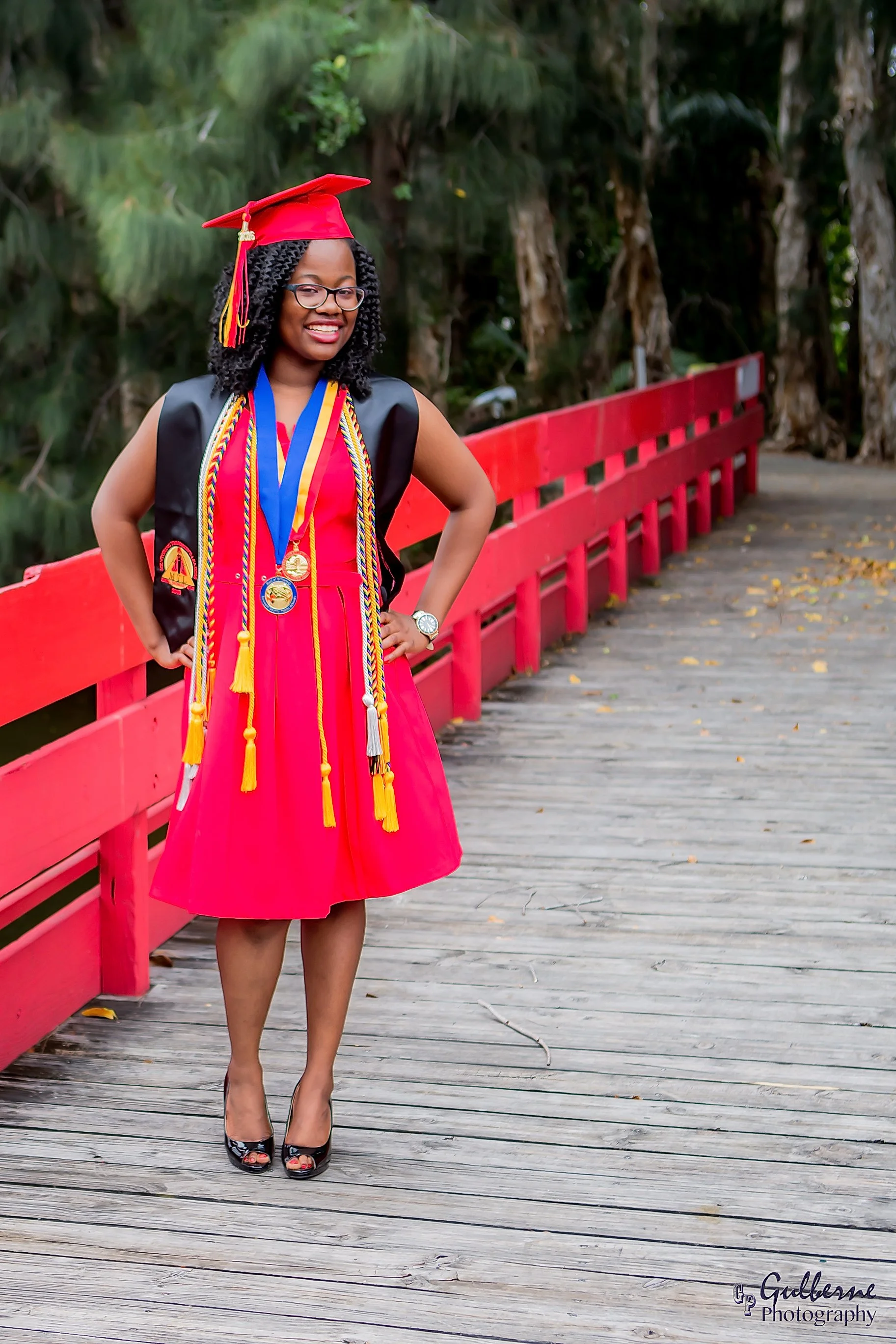 A young woman in a red graduation cap and dress, wearing multiple honor cords and medals, standing on a wooden bridge with a red railing, surrounded by trees.