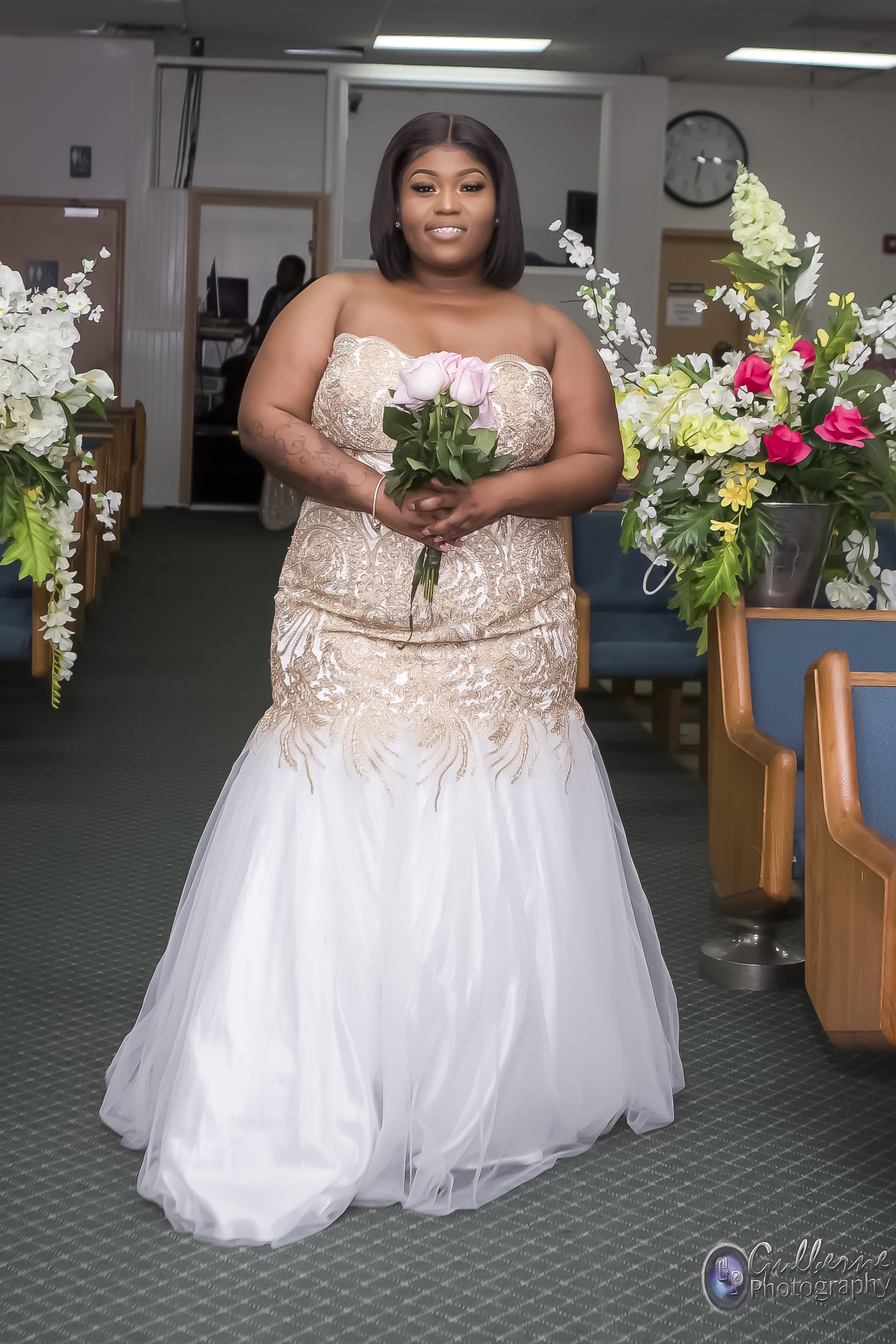 A woman in a strapless, ornate wedding gown holding a bouquet of pink flowers standing in a church decorated with floral arrangements.