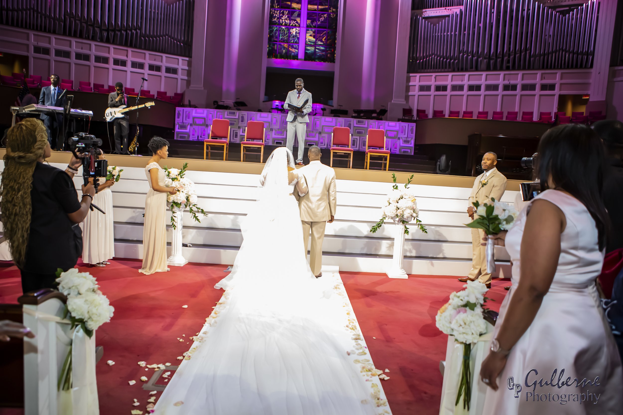 A wedding ceremony taking place in a church with a bride and groom standing at the altar, surrounded by bridesmaids and groomsmen, with clergy reading from a book. There are musicians on the stage and guests seated in pews, holding flowers.