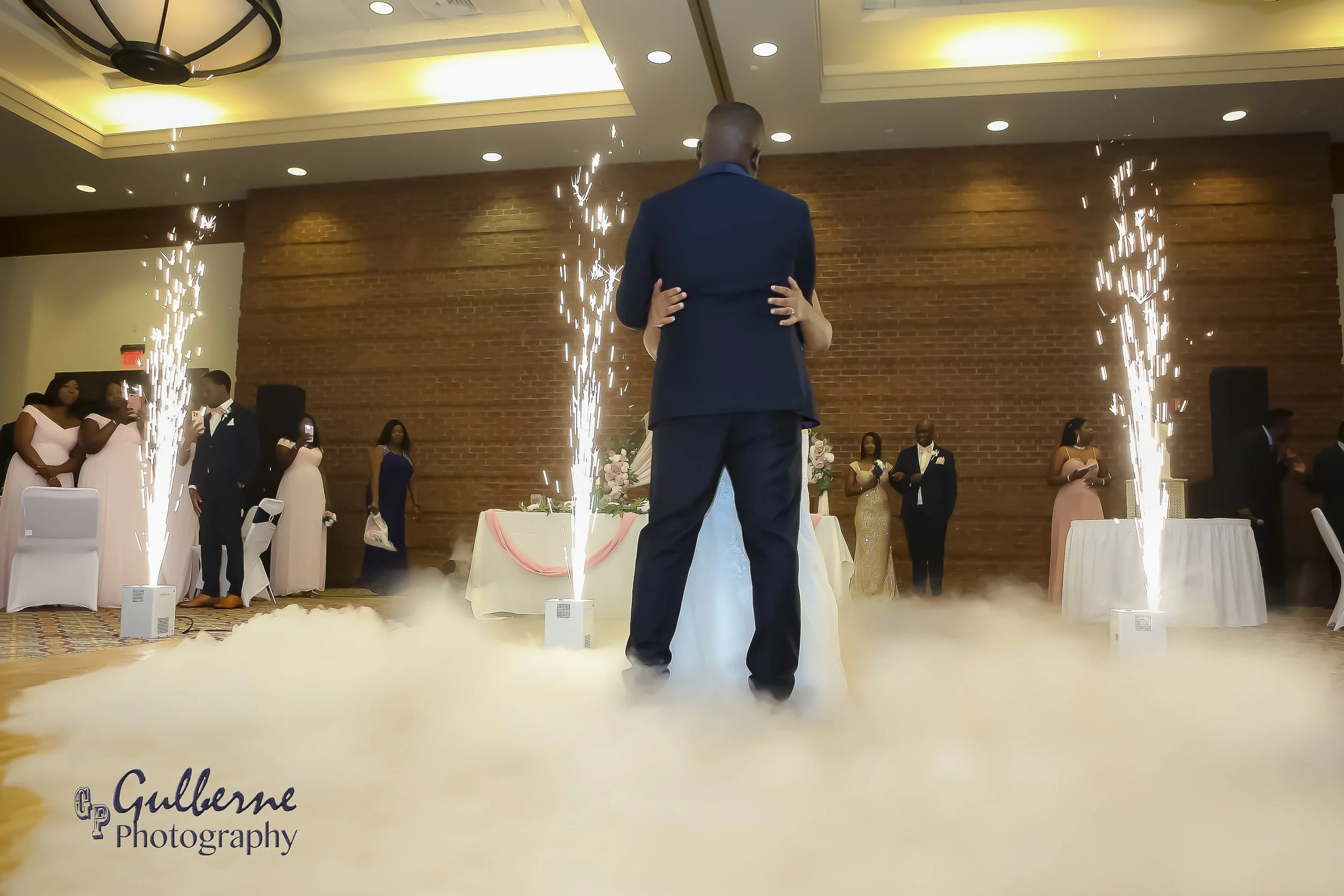 A bride and groom dance at their wedding reception, surrounded by guests. Fireworks are set off on the dance floor with a brick wall background and soft yellow lighting overhead.
