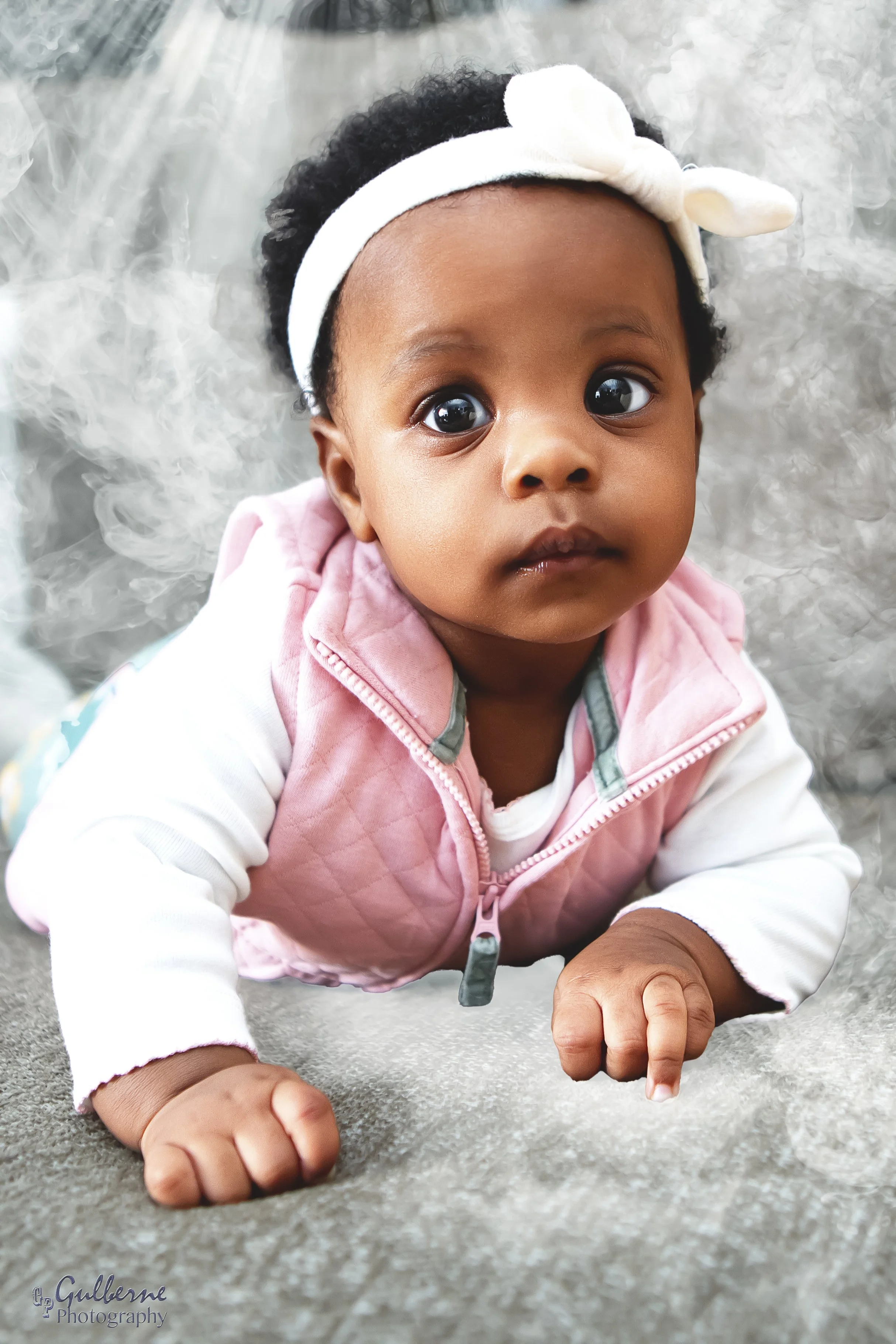 Close-up of a young child with dark curly hair, wearing a white headband with a bow, lying on a fluffy gray surface, looking directly at the camera.